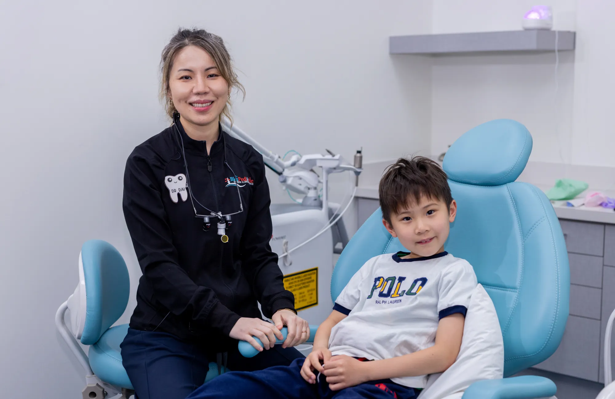 A female dentist in black uniform smiling next to a young boy sitting in a blue dental chair in a dental clinic.