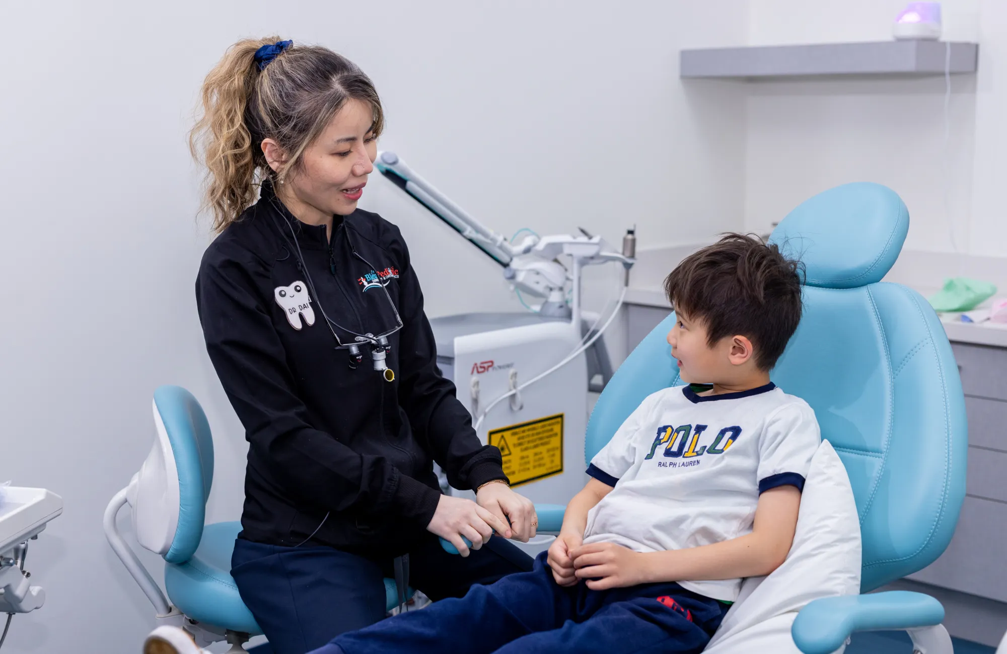 Dentist talking reassuringly with a young boy sitting in a blue dental chair.