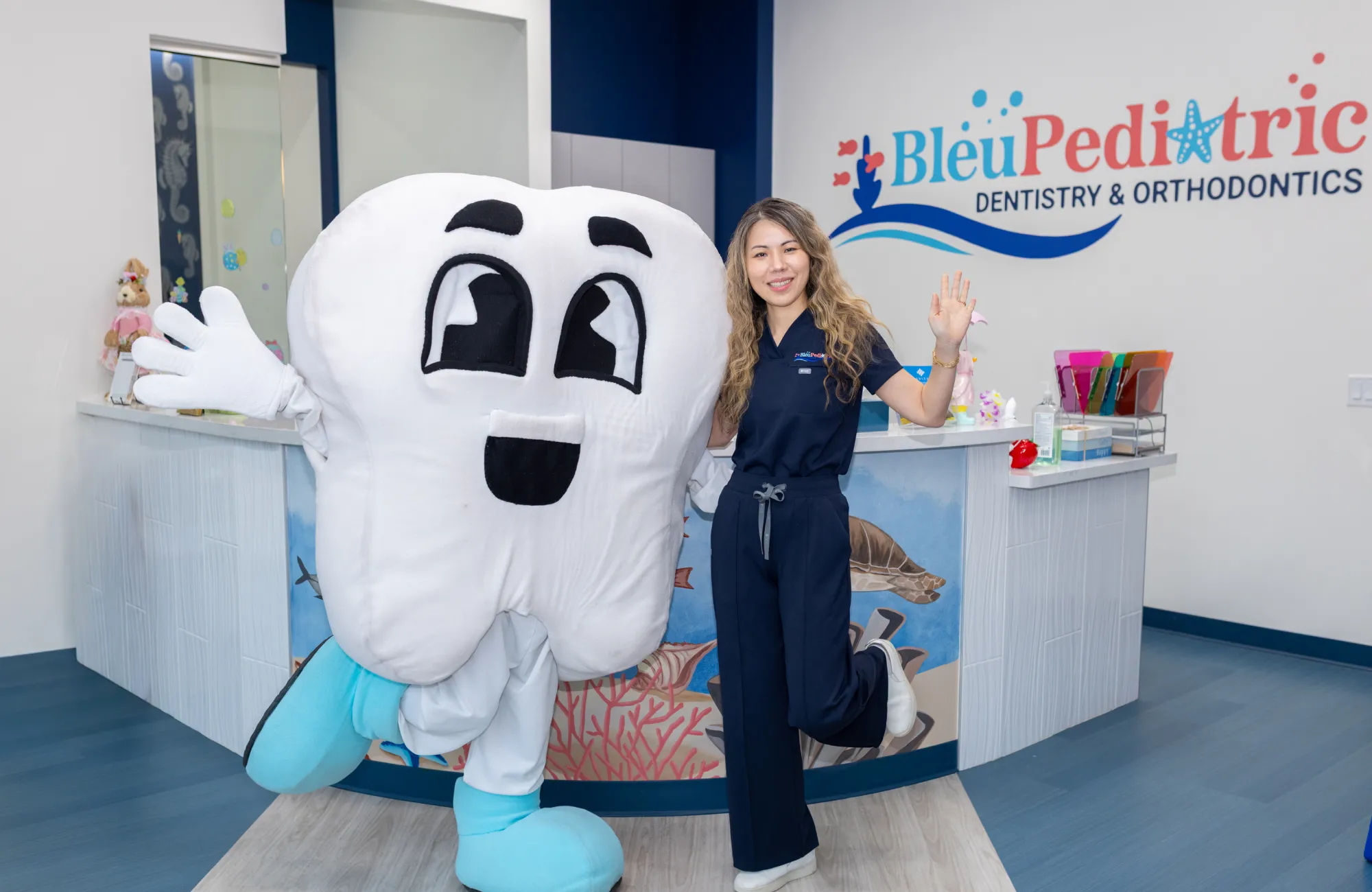 Smiling woman in navy scrubs stands on one leg next to a person in a large smiling tooth costume inside Bleu Pediatric Dentistry & Orthodontics clinic.