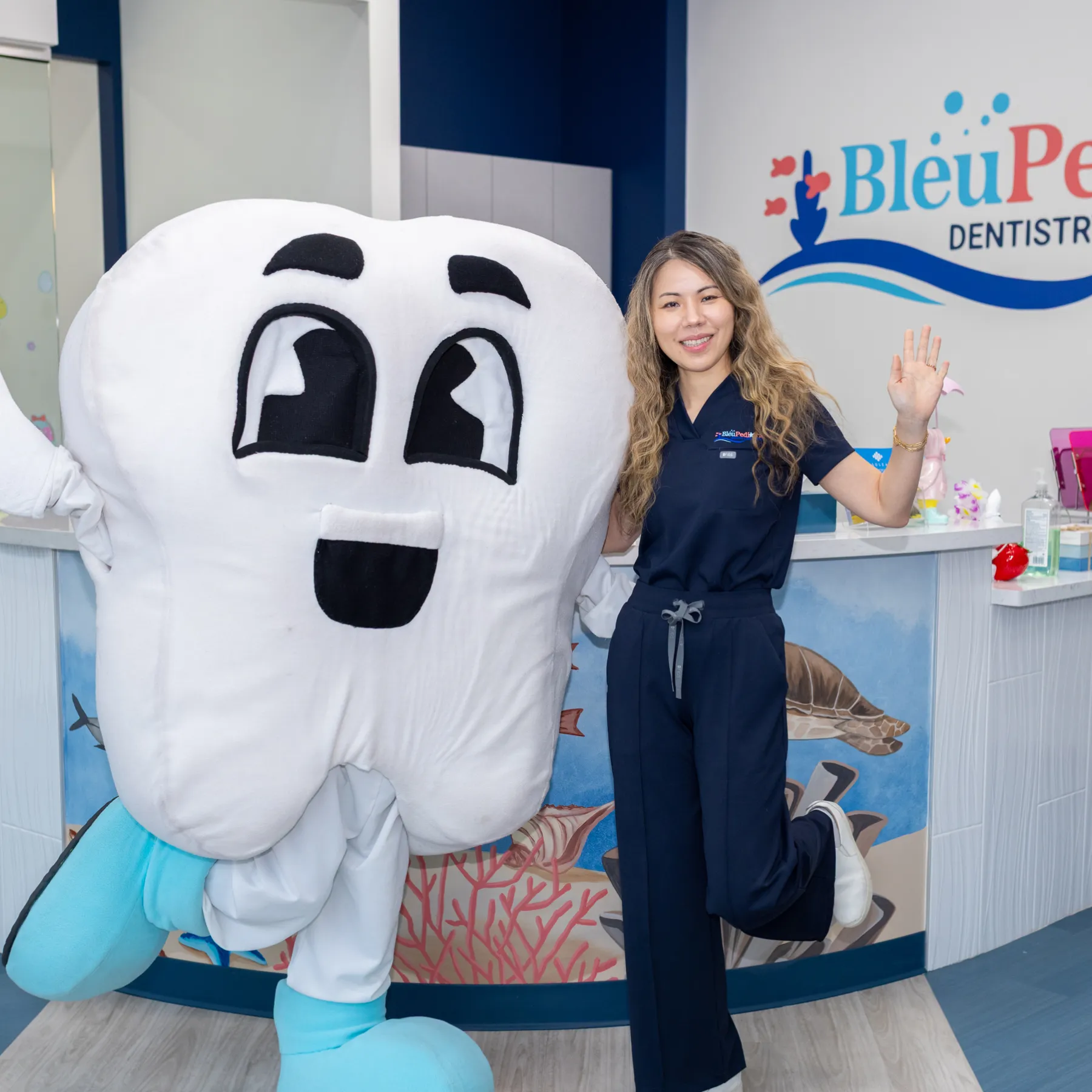 Woman in navy scrubs standing next to a large smiling tooth mascot inside a pediatric dental office.
