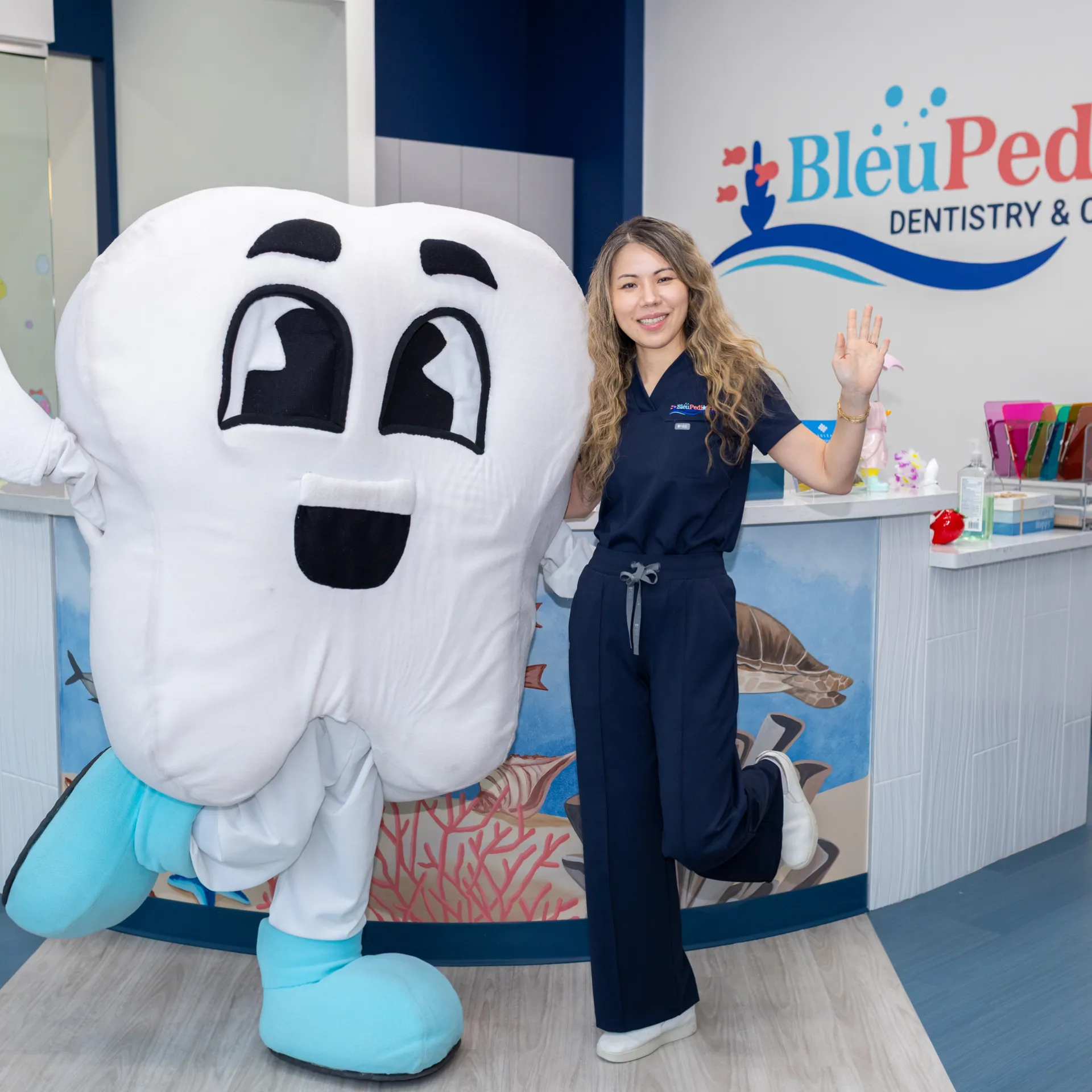 Woman in navy dental scrubs posing with a person in a large smiling tooth mascot costume inside a pediatric dentistry office.