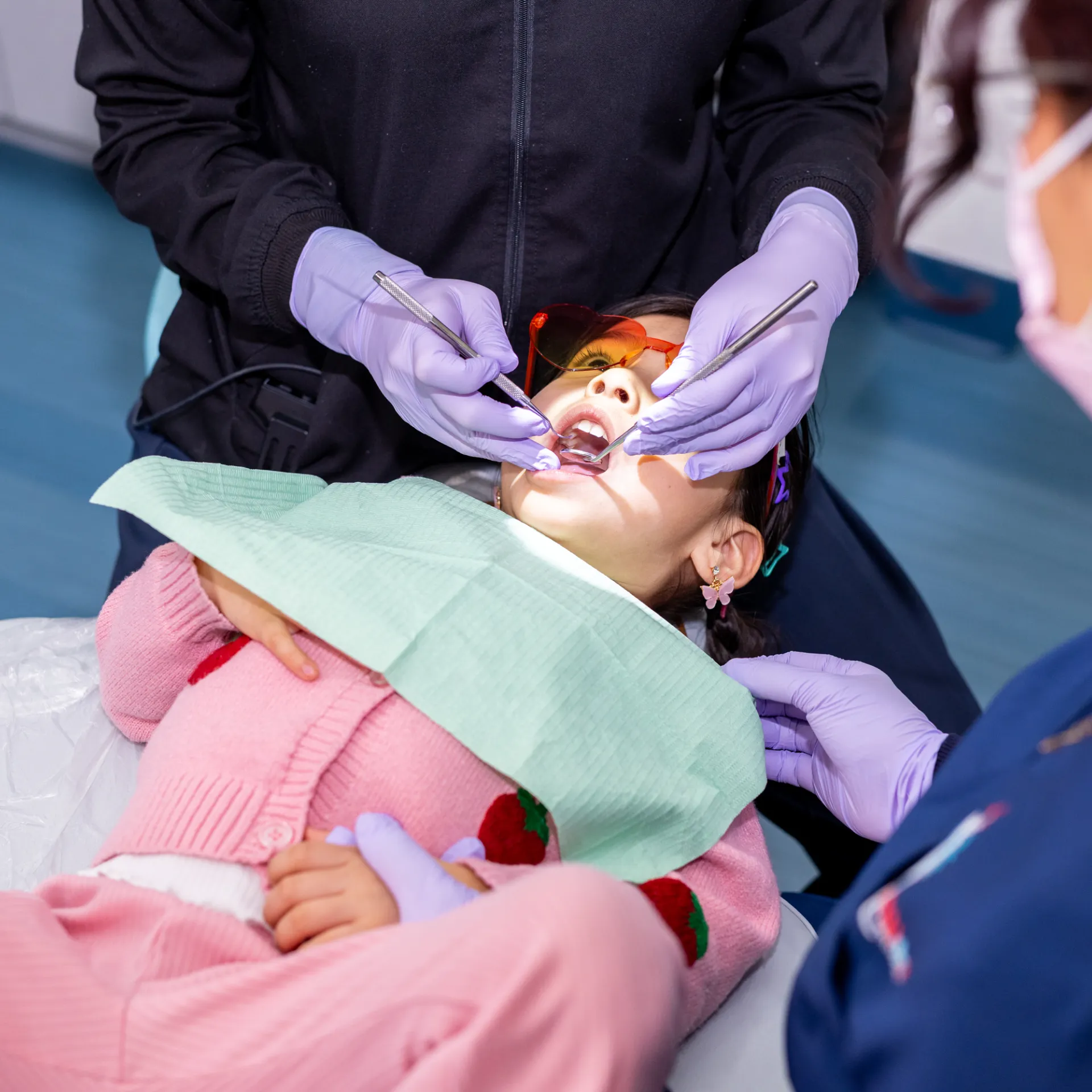 Child wearing orange protective glasses receiving a dental checkup from a dentist in purple gloves.