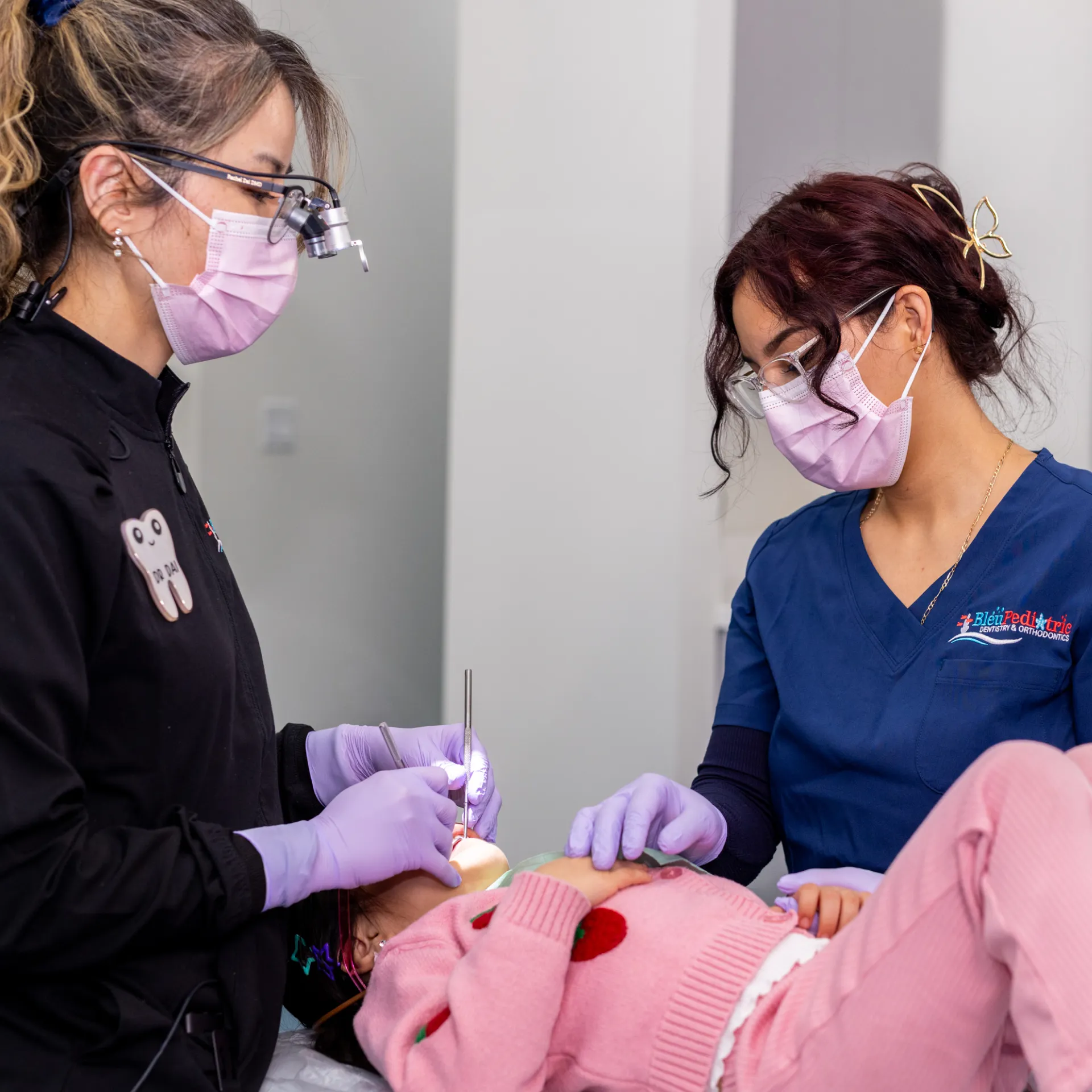 Dental professional performing a procedure on a child patient while another staff member gently holds the child’s hand.