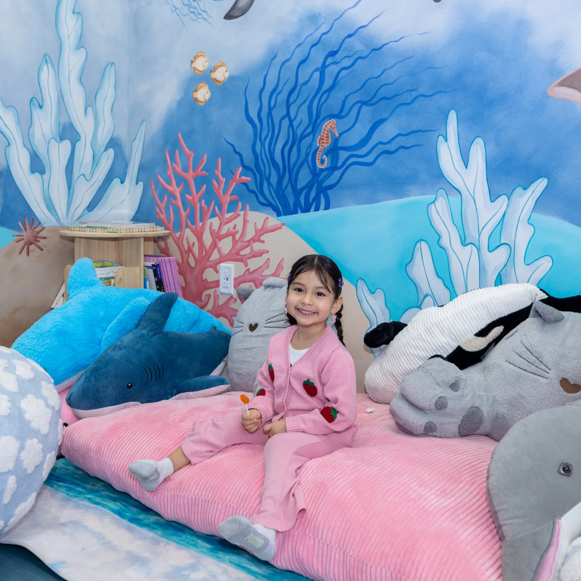 Smiling young girl in pink outfit sitting on a pink cushion surrounded by large plush sea animals with an underwater mural background.