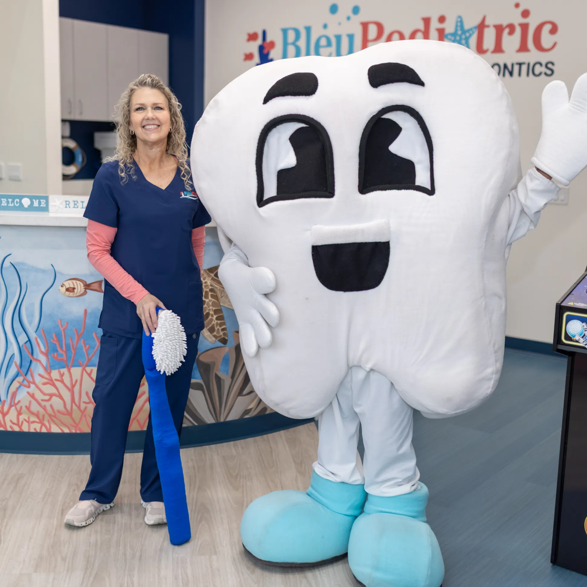 Dental clinic staff member in scrubs standing next to a person in a large tooth mascot costume holding a giant blue toothbrush.