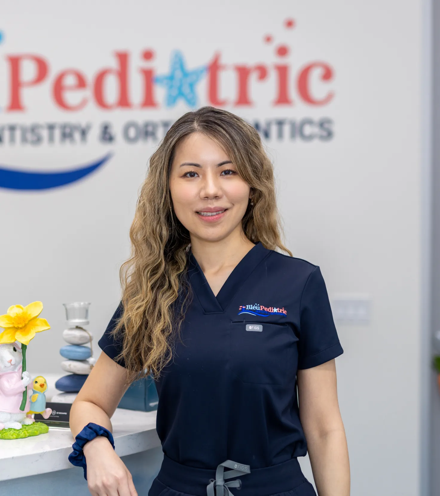 Female pediatric dentist wearing navy scrubs standing with hand on counter in dental office.