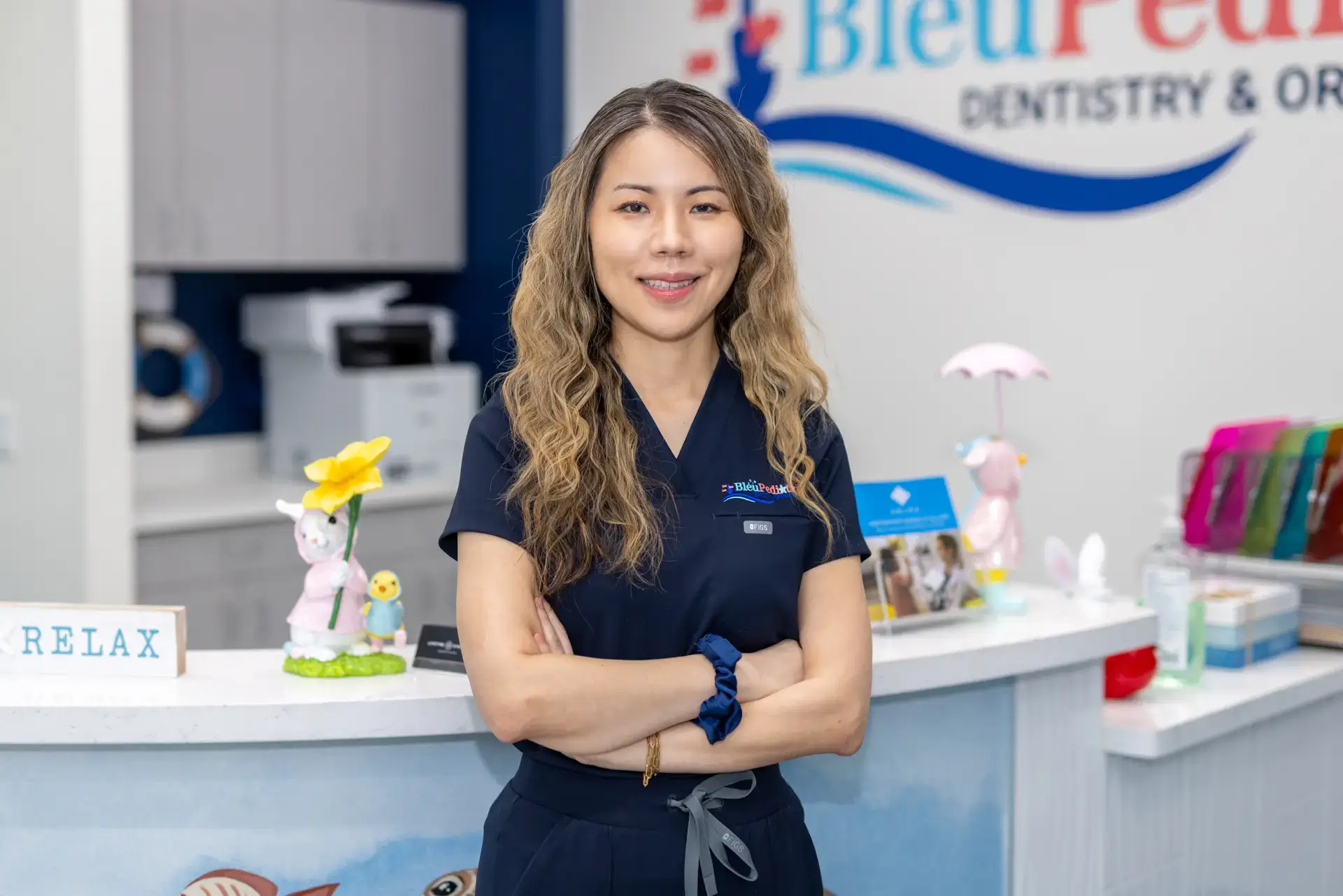 Smiling female dental professional with long curly hair standing with arms crossed in a pediatric dental office reception area.