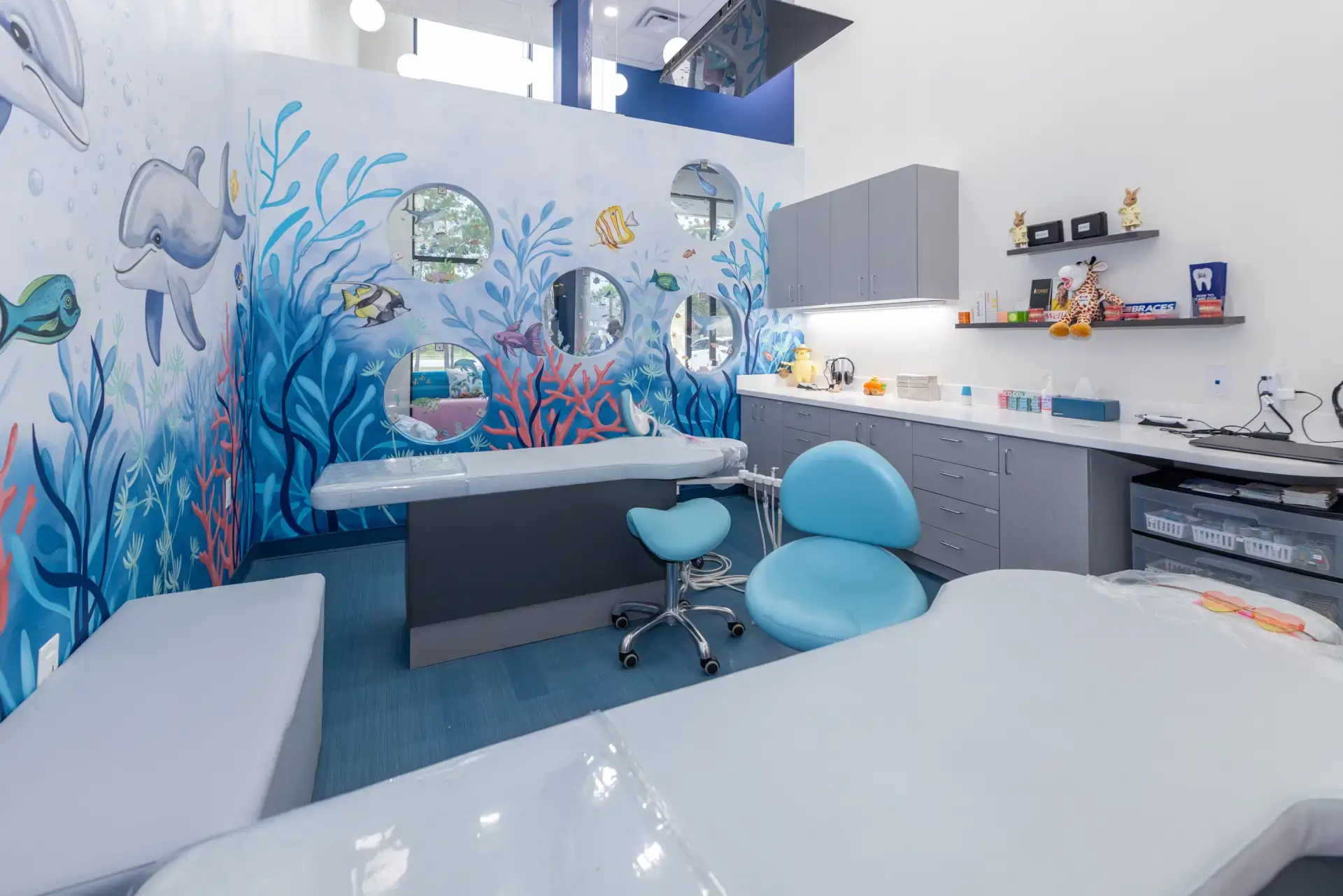 Bright pediatric dental office with underwater-themed wall mural featuring fish and dolphins, blue dental chair, and gray cabinets.