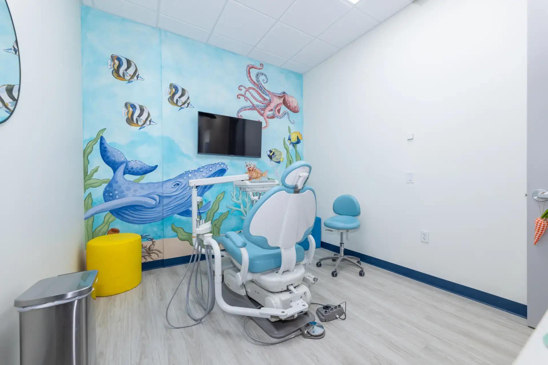 Pediatric dental exam room with ocean-themed wall mural featuring a whale, fish, and octopus, a dental chair, and a TV.