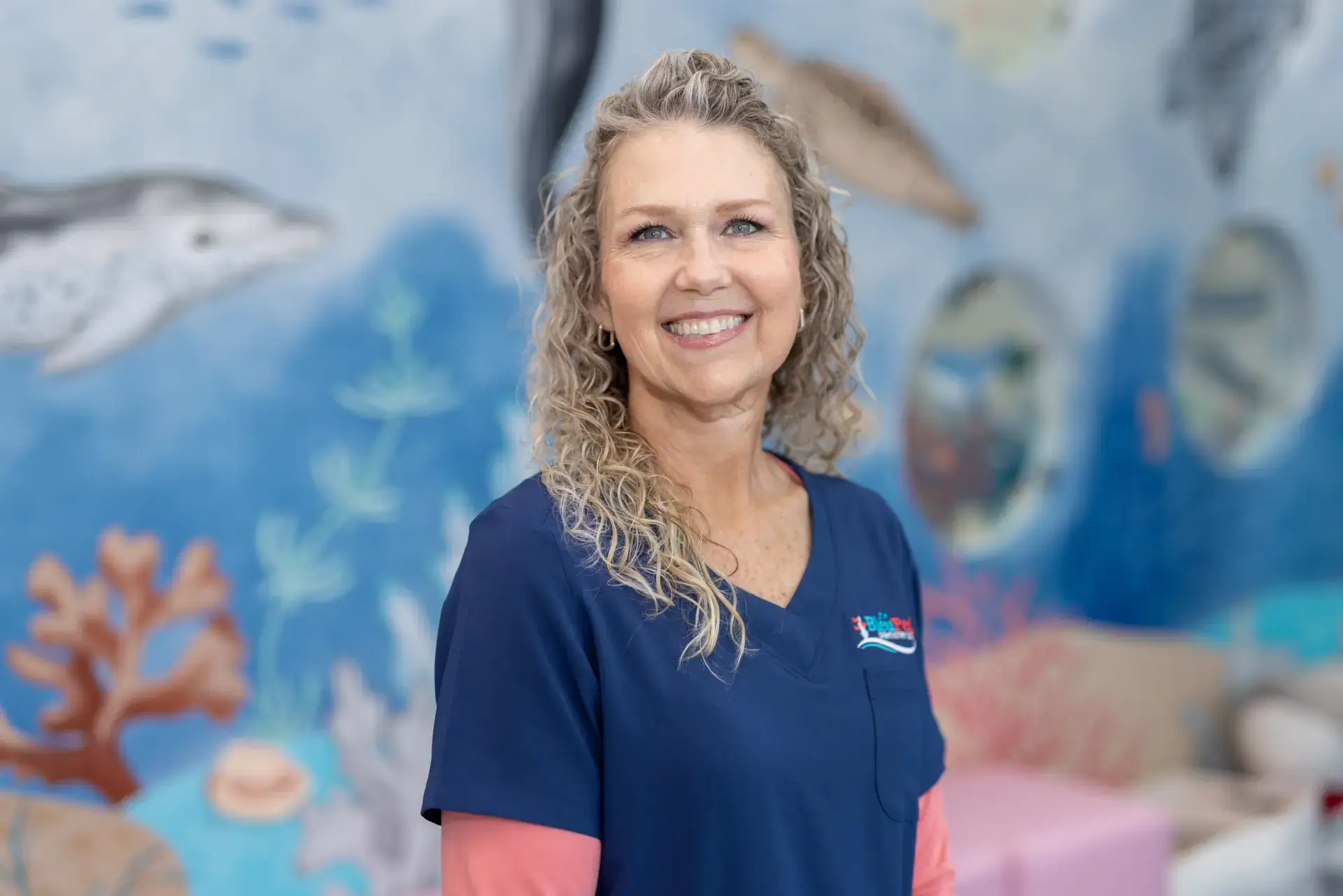 Smiling woman with curly blonde hair in a navy blue medical scrub top standing in front of a colorful underwater-themed mural.