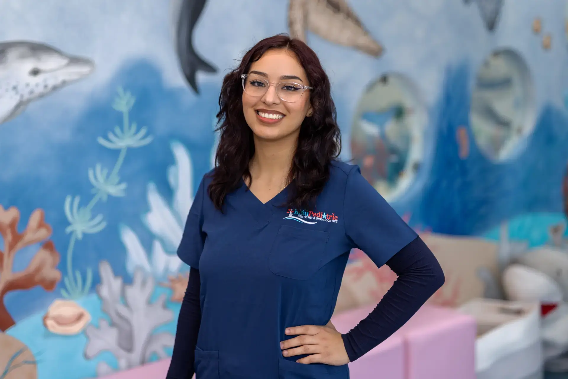 Smiling woman in blue medical scrubs with transparent glasses standing in front of a marine life mural.