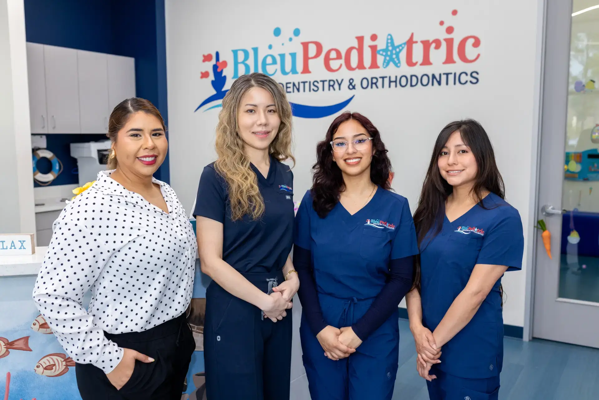Four smiling women posing inside Bleu Pediatric Dentistry & Orthodontics clinic, three in navy scrubs and one in white blouse with black polka dots.