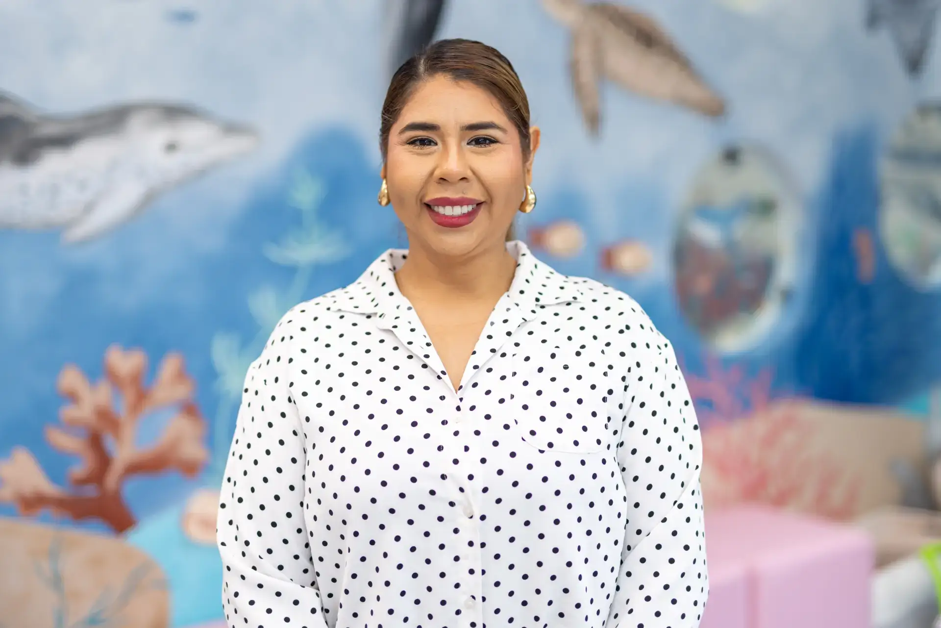 Smiling woman with gold hoop earrings wearing a white blouse with black polka dots in front of a blue underwater-themed mural.