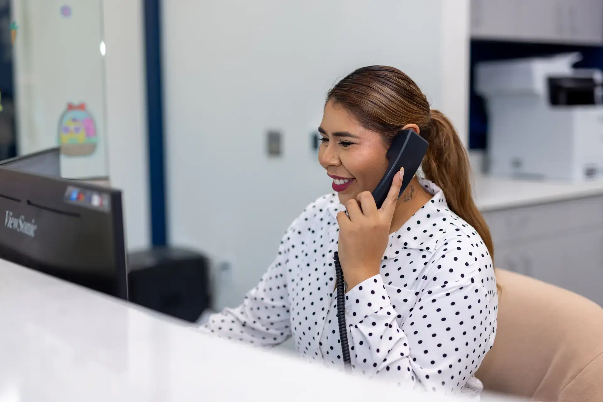 Smiling woman with a ponytail wearing a white polka dot shirt, talking on a desk phone in an office setting.