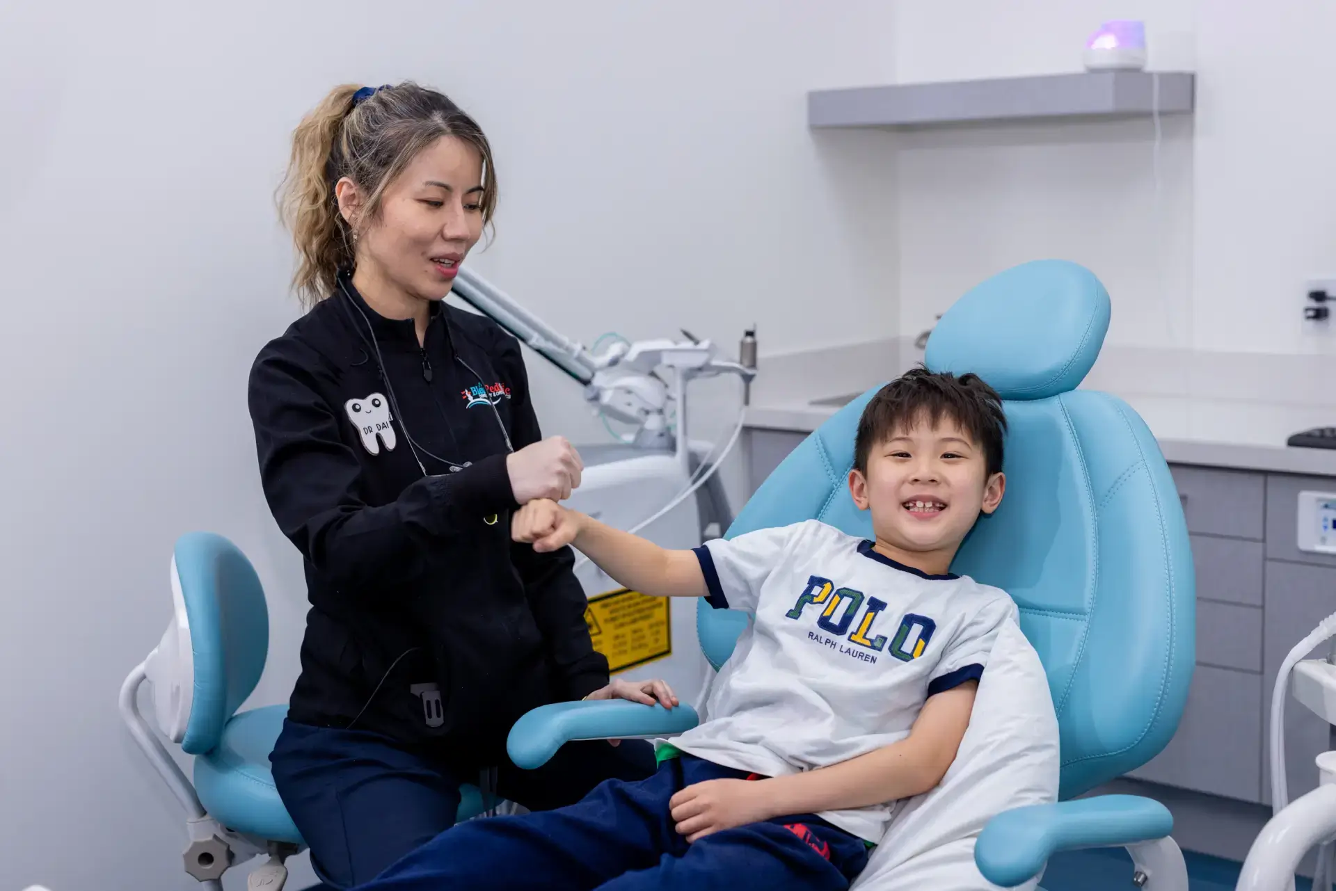 A female dentist fist bumping a smiling young boy sitting in a blue dental chair in a clinic.