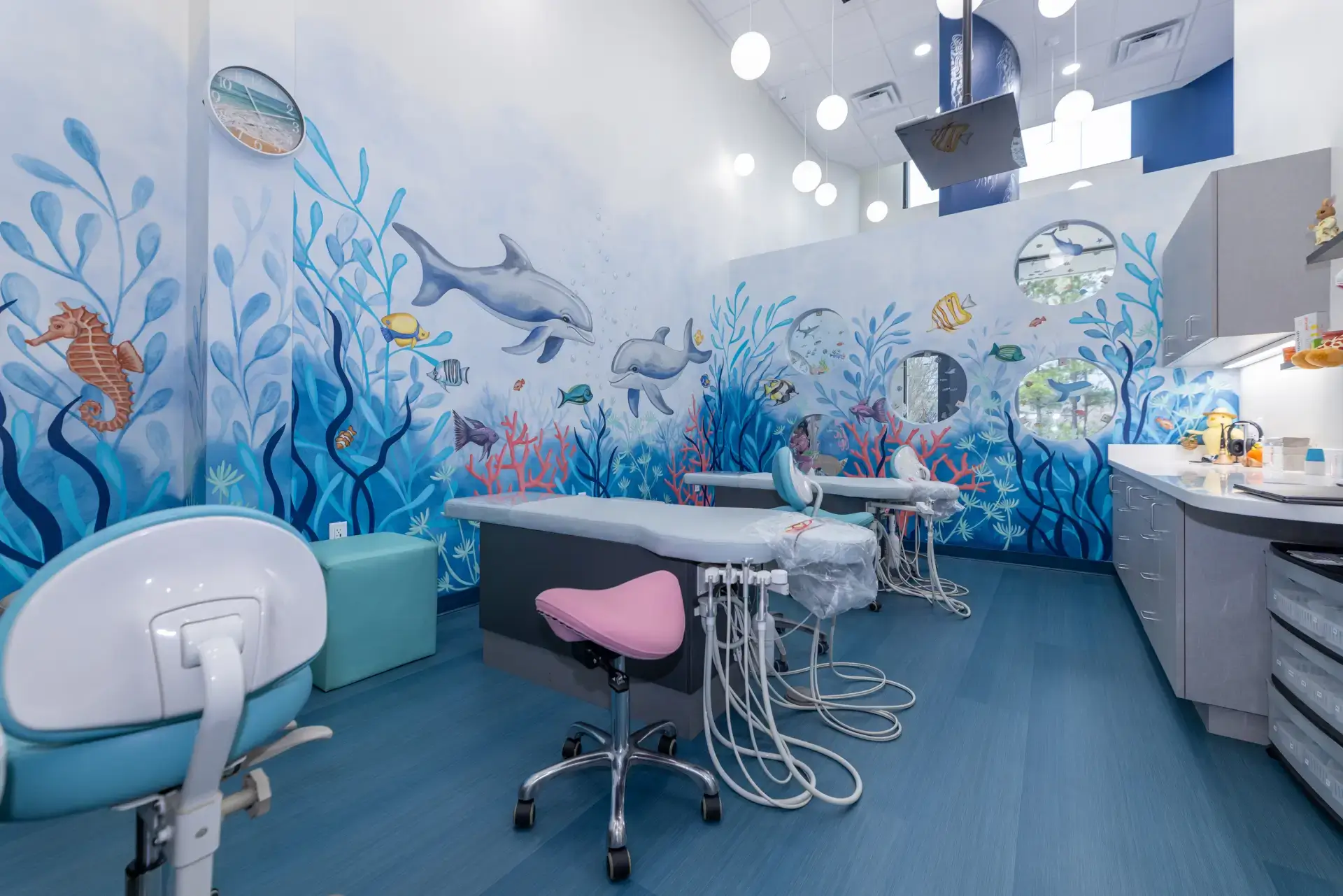 Pediatric dental clinic room with aquatic-themed wall murals featuring dolphins, fish, and sea plants, dental chairs, and equipment.