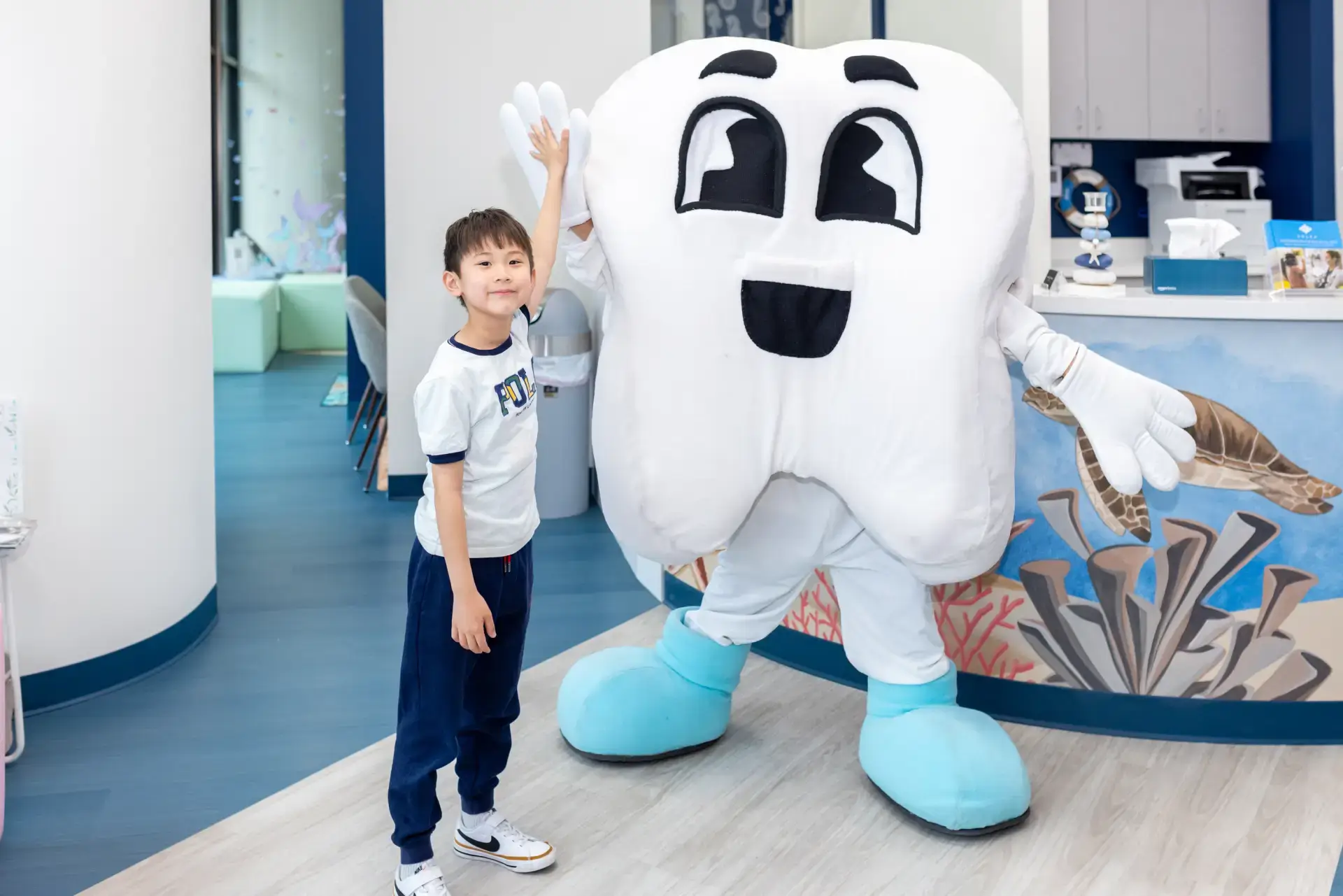Child giving a high five to a person in a large smiling tooth costume in a dental office.