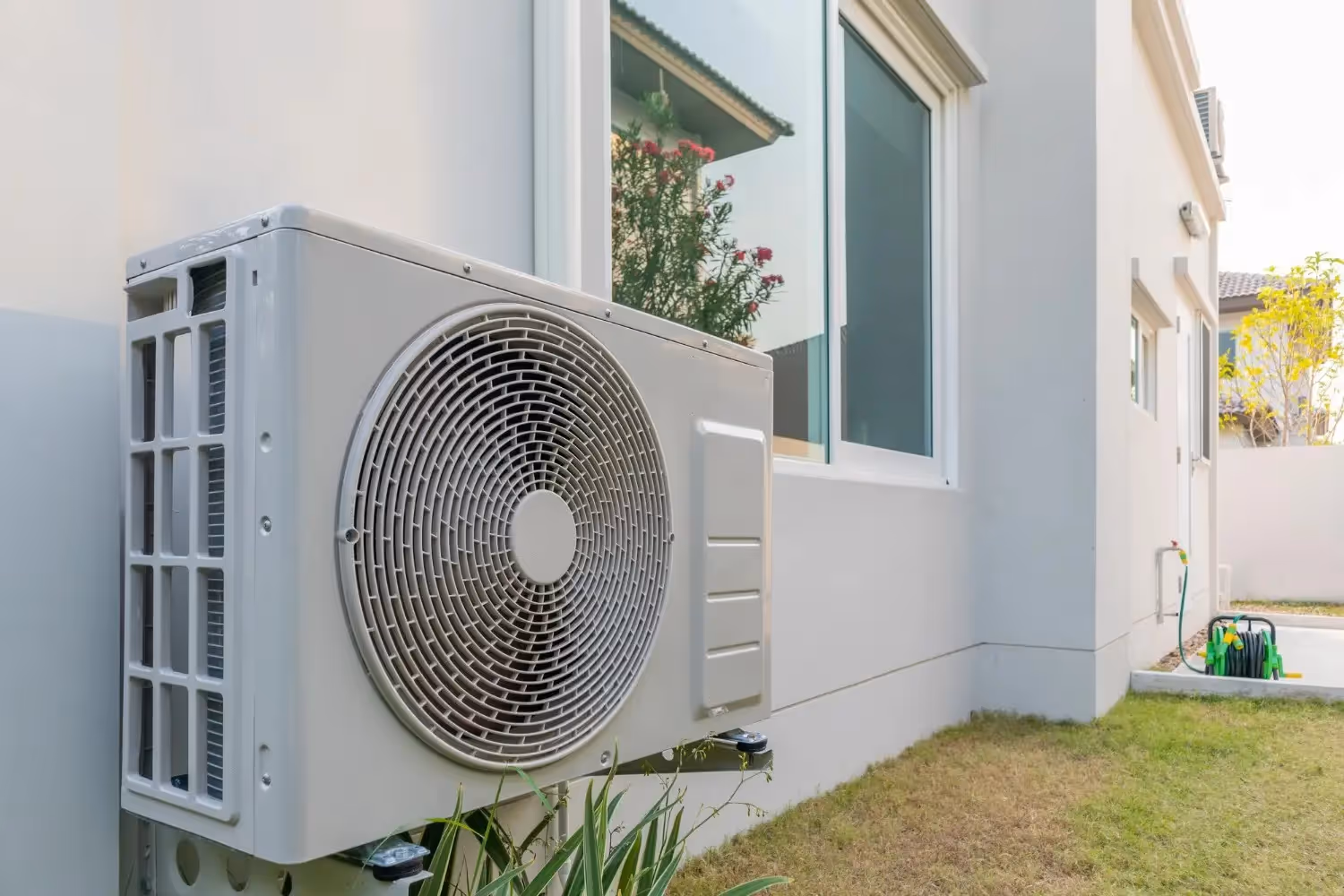 Air conditioning unit mounted on white wall near flowering plants and window
