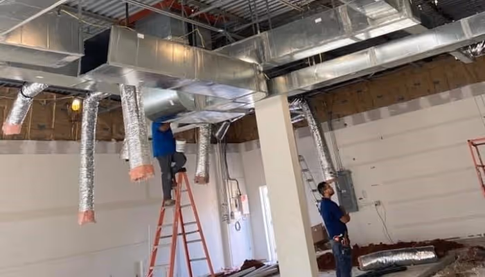 Workers installing industrial HVAC ductwork on ladders in an unfinished building