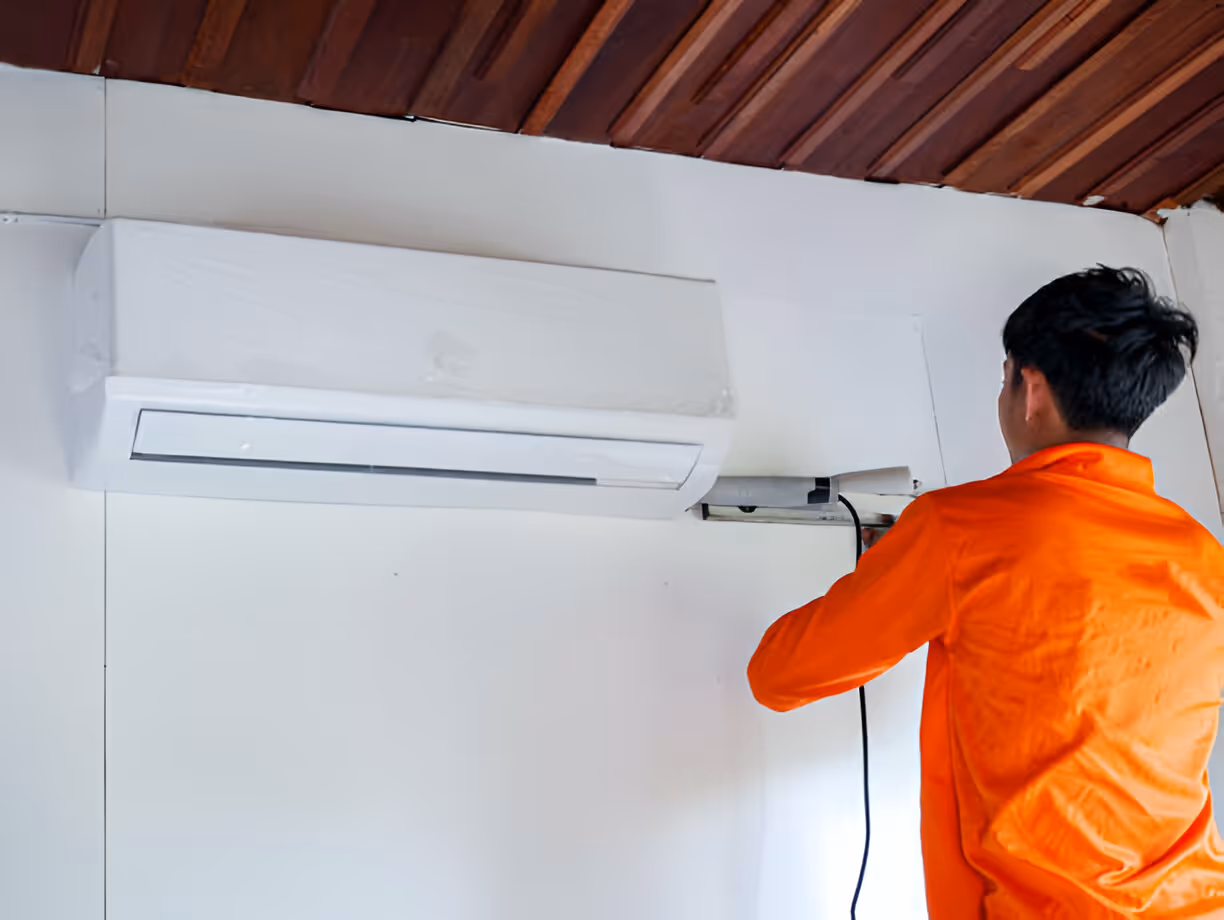 Technician in orange shirt installing or repairing a white wall-mounted air conditioner