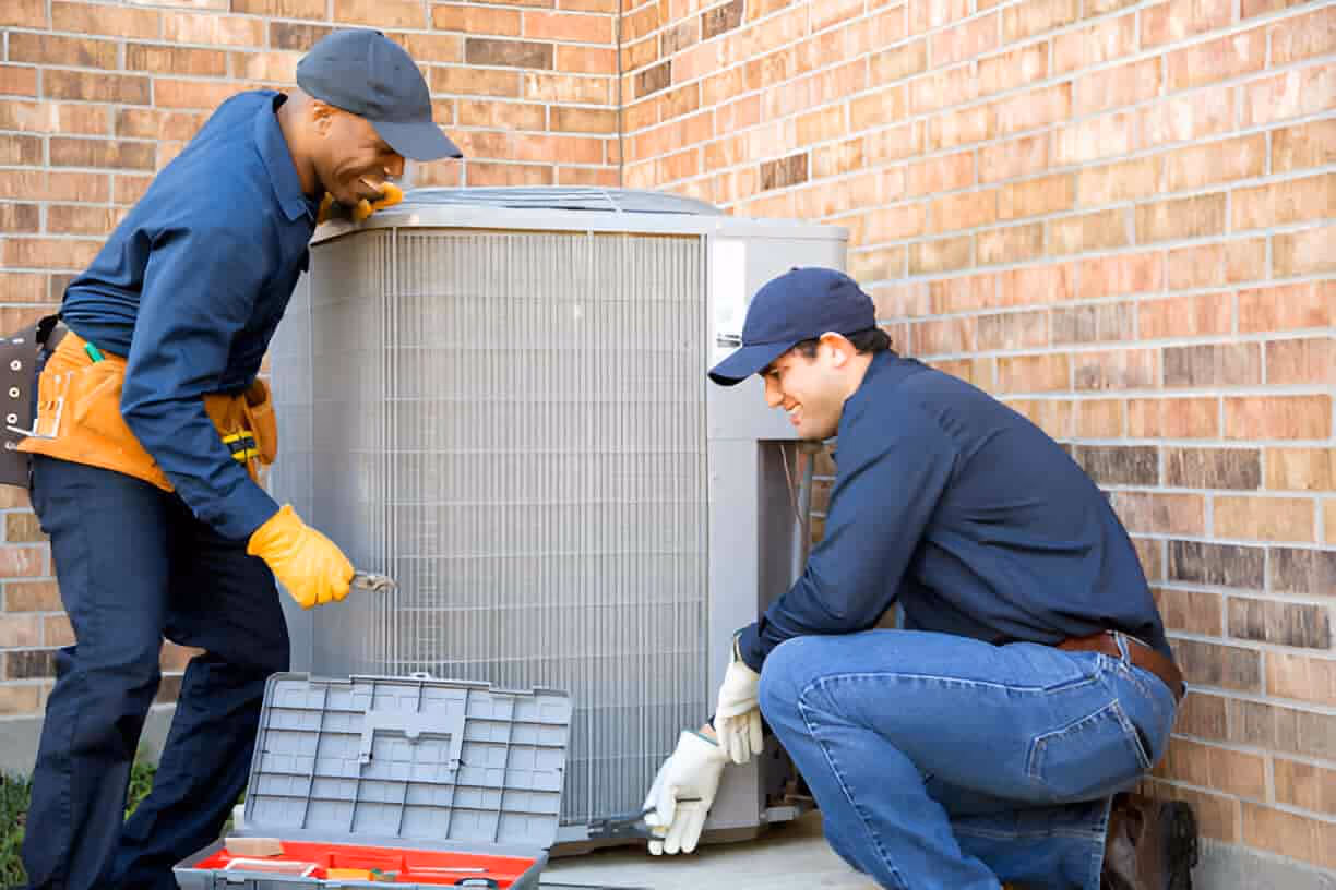 Two technicians repairing an air conditioning unit outside a brick building