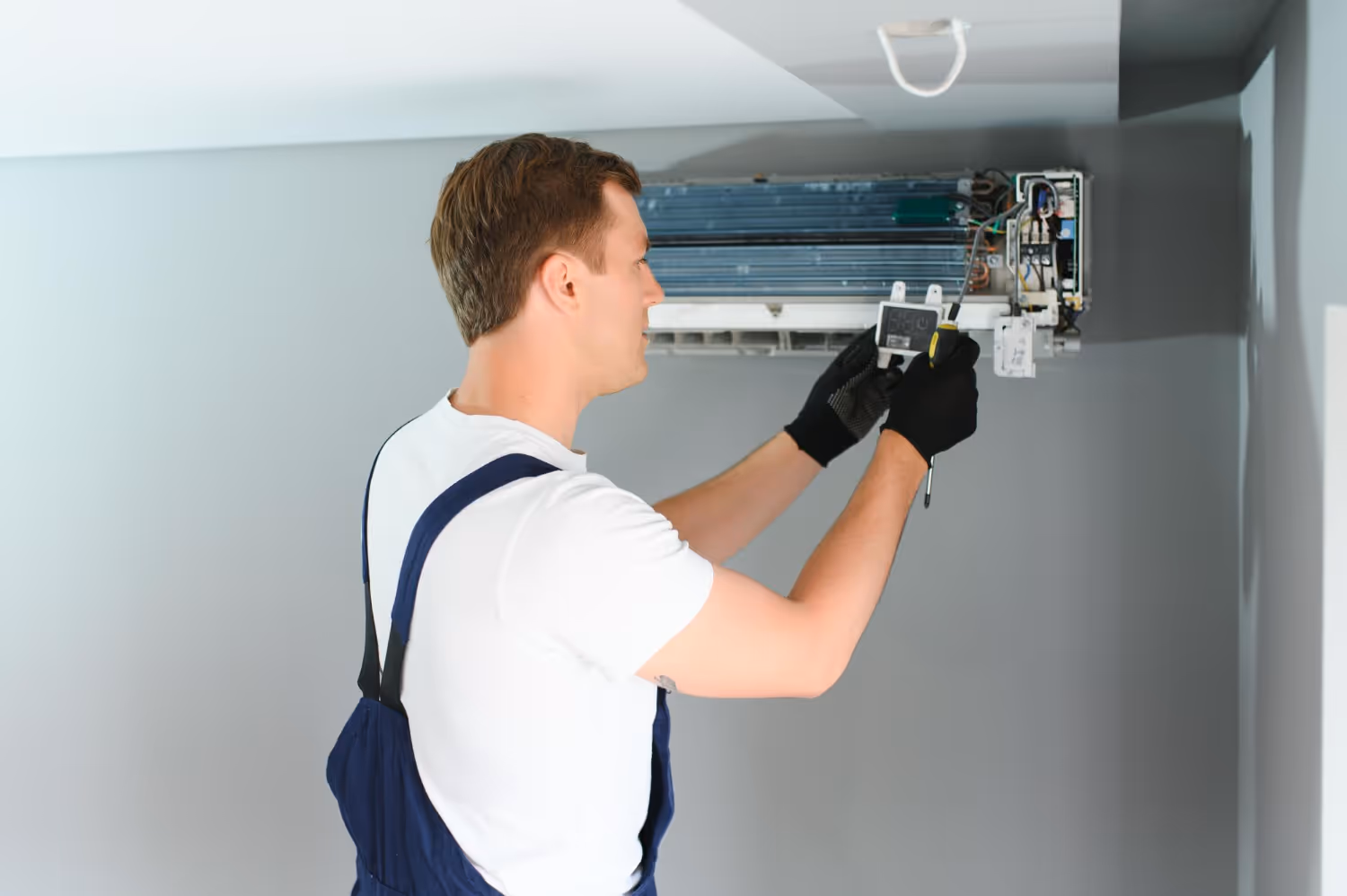 Technician in white shirt and blue overalls repairing air conditioning unit