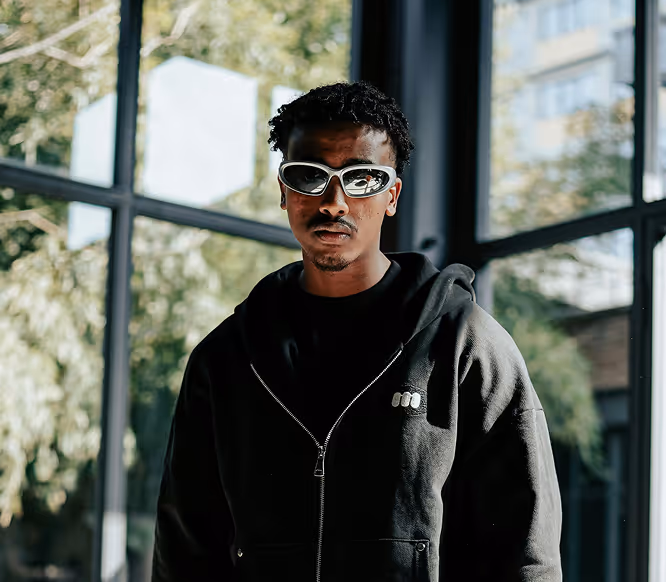 Trendy man in dark streetwear and bold sunglasses posing indoors, representing edgy influencer style powered by Kicksti visibility.