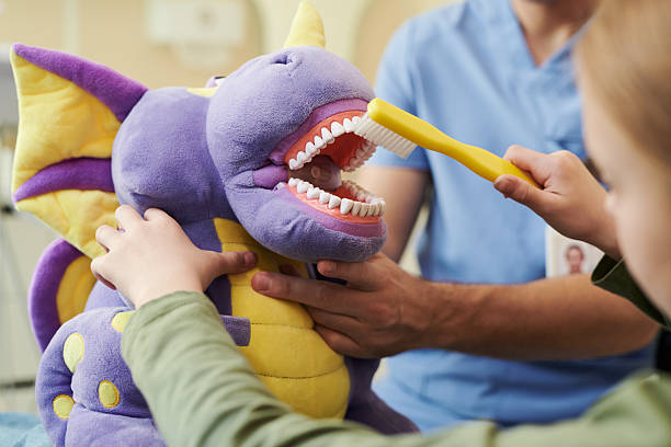 Child Brushing Teeth of Plush Dragon Toy during Dental Hygiene Demonstration Child brushing teeth of large purple dragon plush toy while adult male dentist supervising, demonstrating dental hygiene technique in dental clinic setting, hands visible Parent and child sitting in a pediatric dental chair, smiling as the dentist explains brushing techniques using a puppet stock pictures, royalty-free photos & images