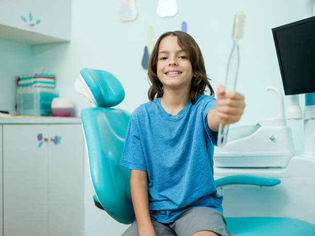 Cute boy holding big toothbrush and smiling at camera A cute latin boy sitting in dental clinic, holding a big toothbrush and smiling at the camera. A young boy sitting in a dental chair, smiling proudly while holding a small toothbrush and wearing a Thanksgiving-themed shirt stock pictures, royalty-free photos & images