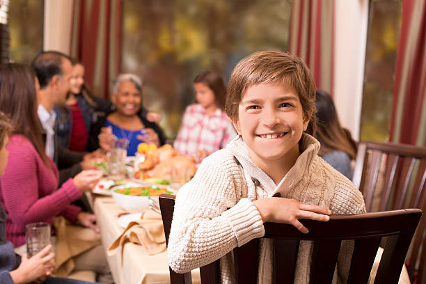 Multi-ethnic family enjoying Thanksgiving dinner at grandmother's home. Multi-ethnic, multi-generation family enjoying Thanksgiving dinner together around the dining table at grandmother's home. Latin descent, African descent, mixed race, and Caucasian family members. Turkey and other food on table. Cute little boy in chair foreground. A smiling boy at a Thanksgiving table holding a small toothbrush next to a plate of pumpkin pie, warm autumn lighting. stock pictures, royalty-free photos & images