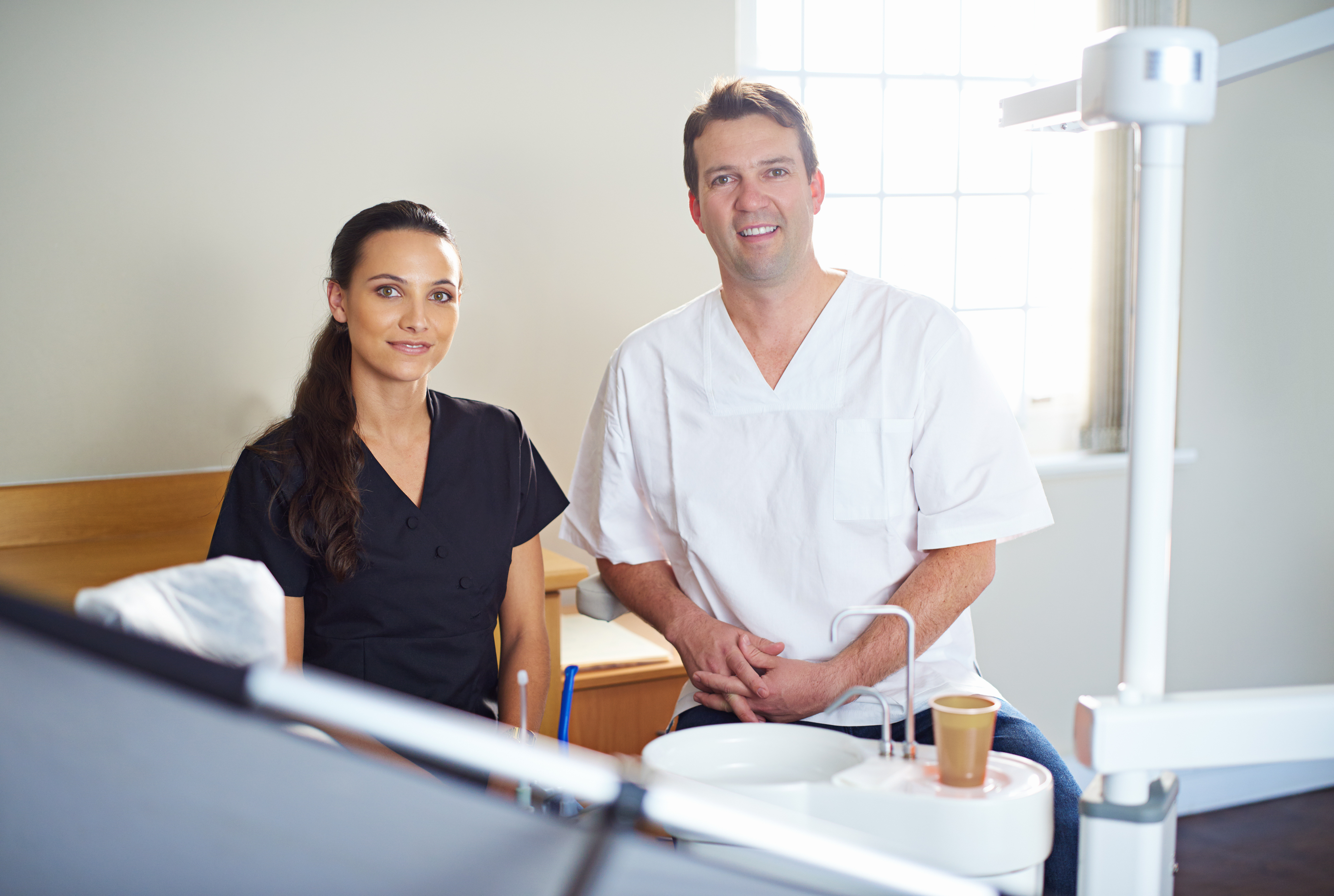 Two healthcare professionals in a dental office, one wearing black scrubs and the other wearing white scrubs, sitting and smiling at the camera.