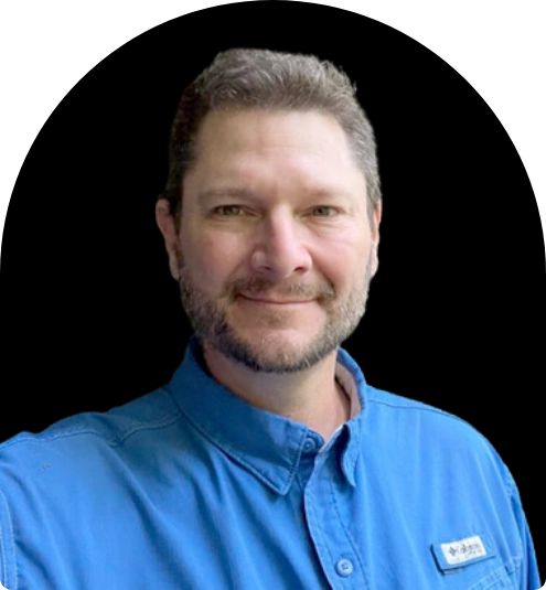 Smiling man with short hair and beard wearing a blue collared shirt against a black background.