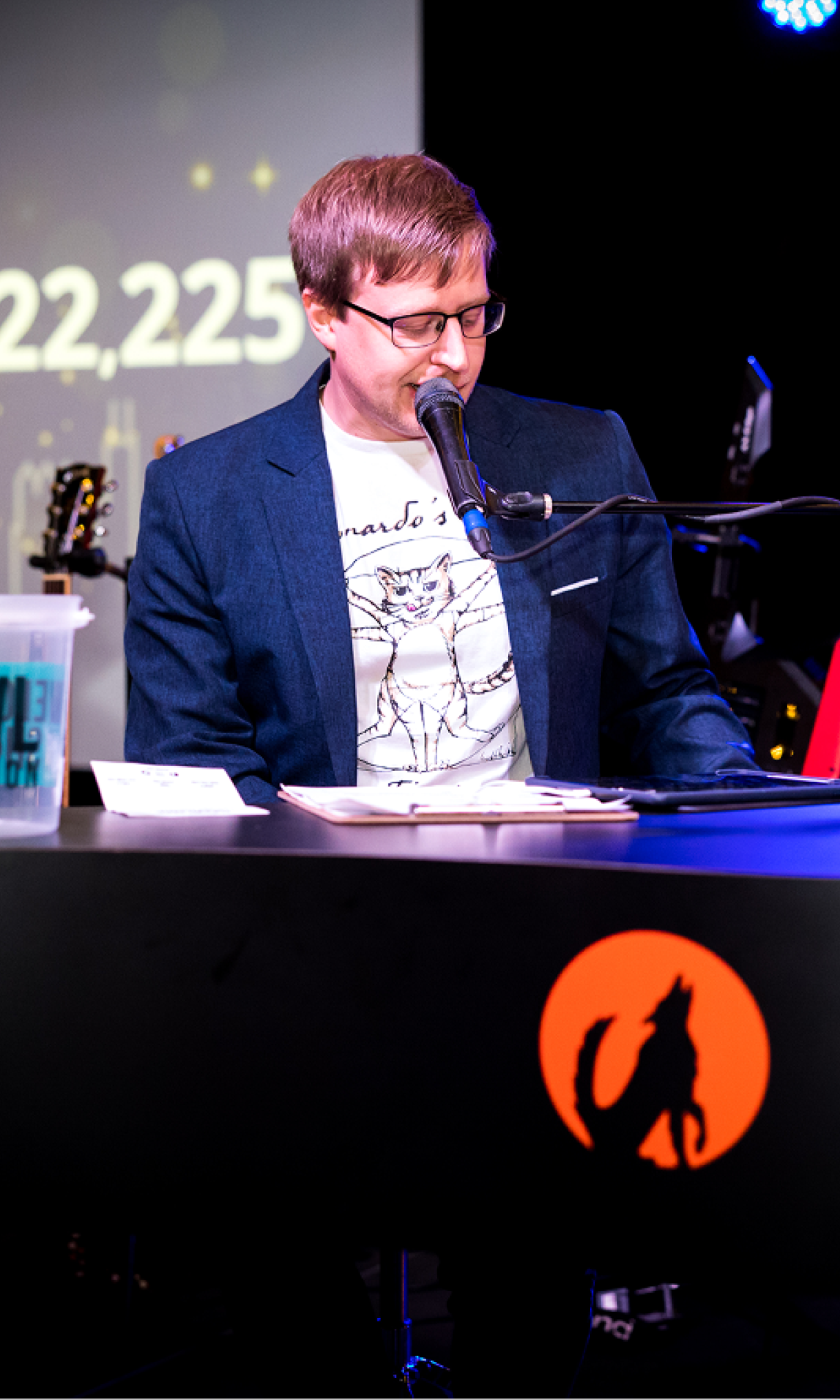 Man playing piano and singing into a microphone, with a fundraising presentation displaying financial information projected in the background.