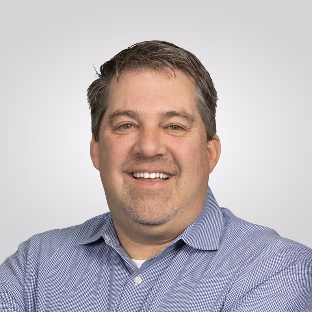 Smiling man with short hair wearing a blue checkered shirt against a plain gray background.