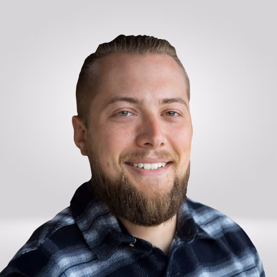 Smiling man with slicked-back hair and a beard wearing a dark blue and gray plaid shirt against a light gray background.