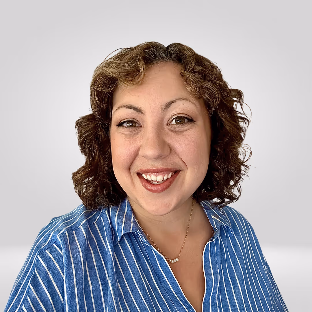 Smiling woman with curly brown hair wearing a blue and white striped shirt and a pearl necklace, against a light gray background.