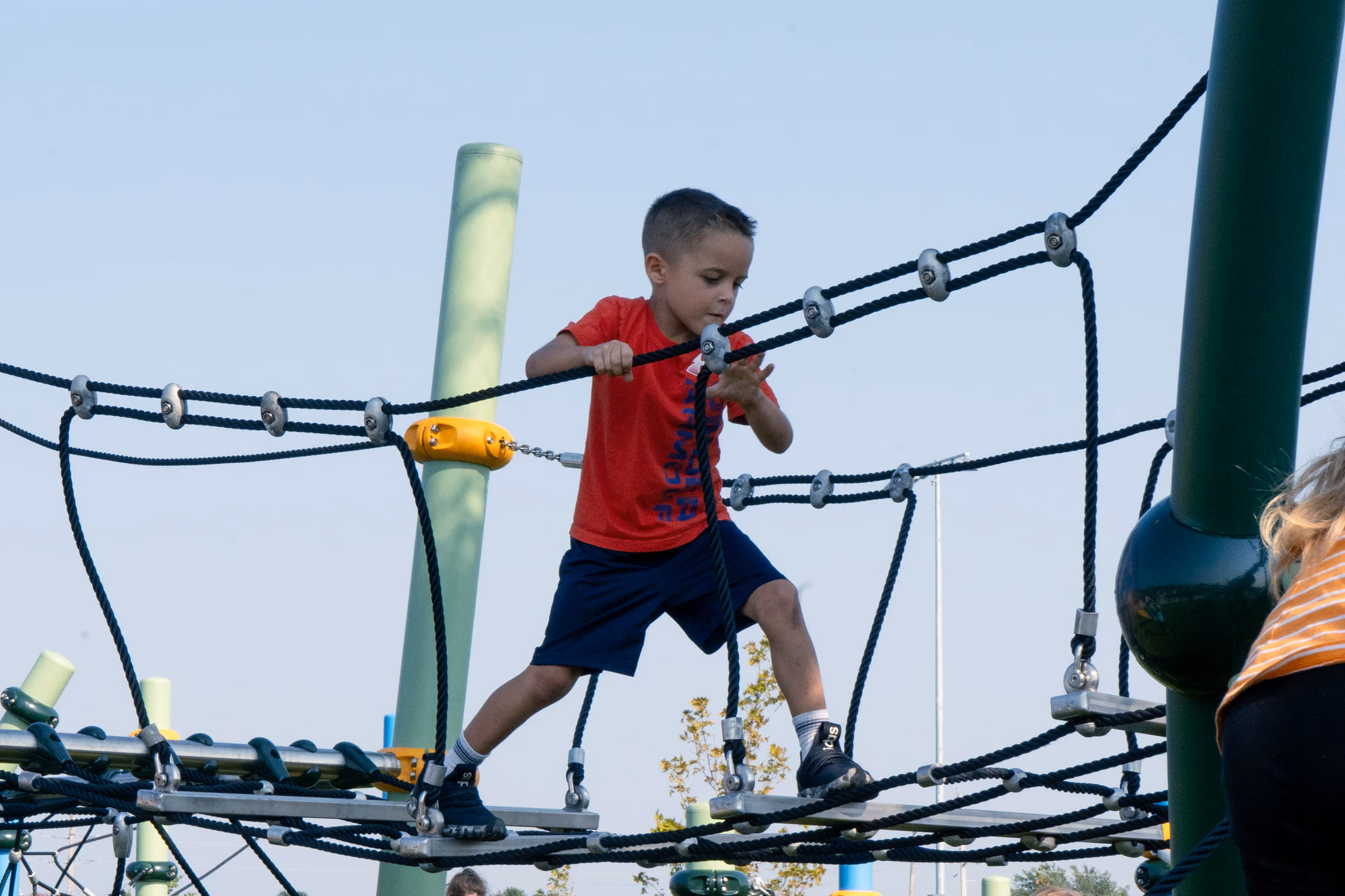 Boy in red shirt and blue shorts climbing on a rope bridge at playground.