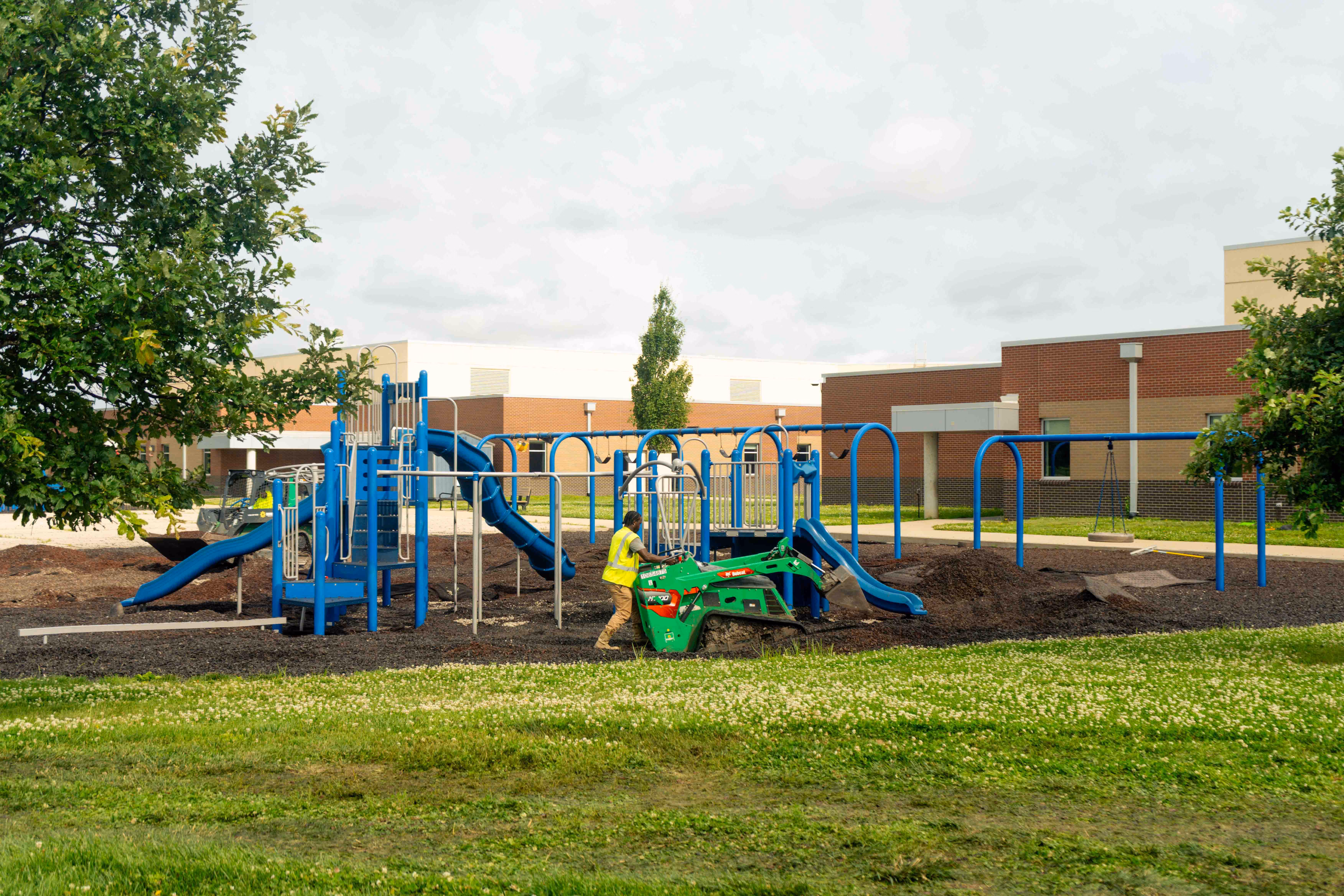 Worker in yellow vest operates small green excavator at playground with blue slides and swings.