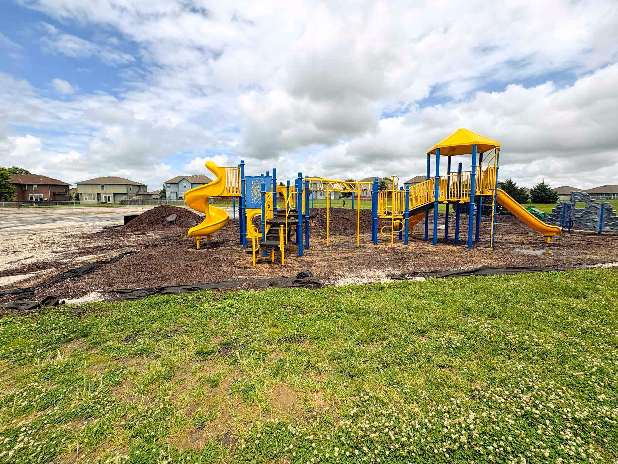 Playground with yellow slides and blue structures on mulch with green grass in foreground and houses in background.