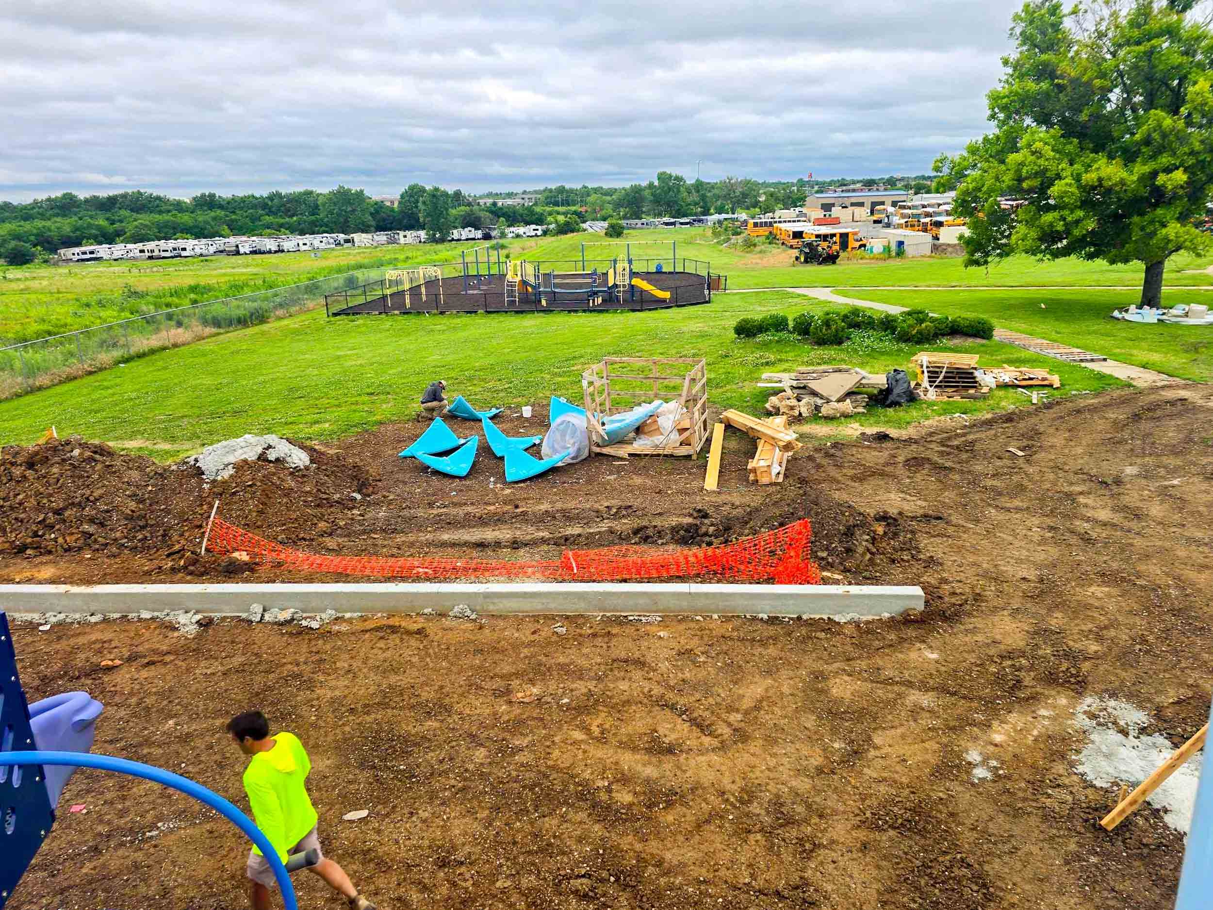 Playground under construction with equipment pieces on dirt and a man walking nearby on a cloudy day.