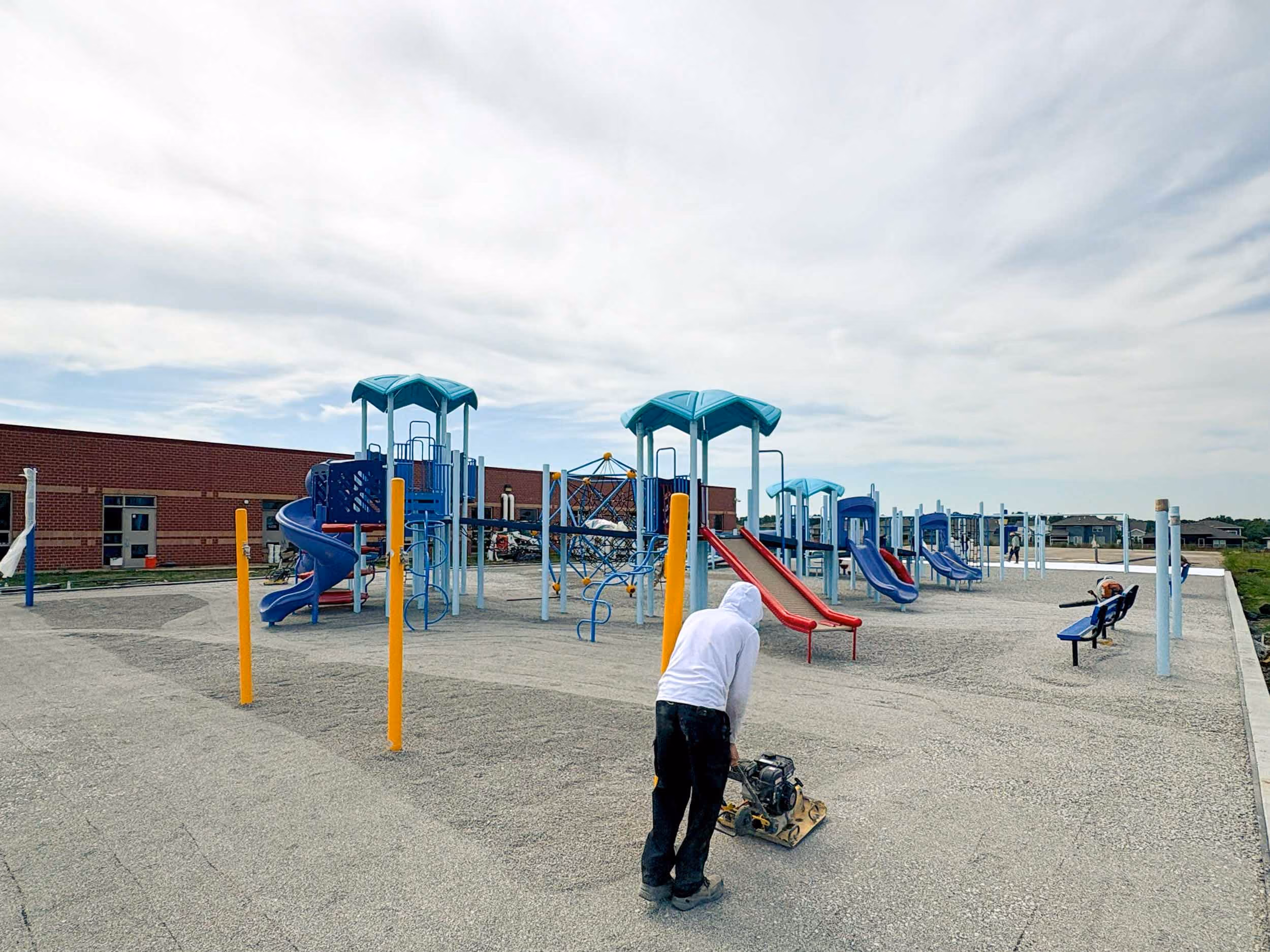 Person operating a compactor machine on a gravel playground near blue and red slides and climbing structures.
