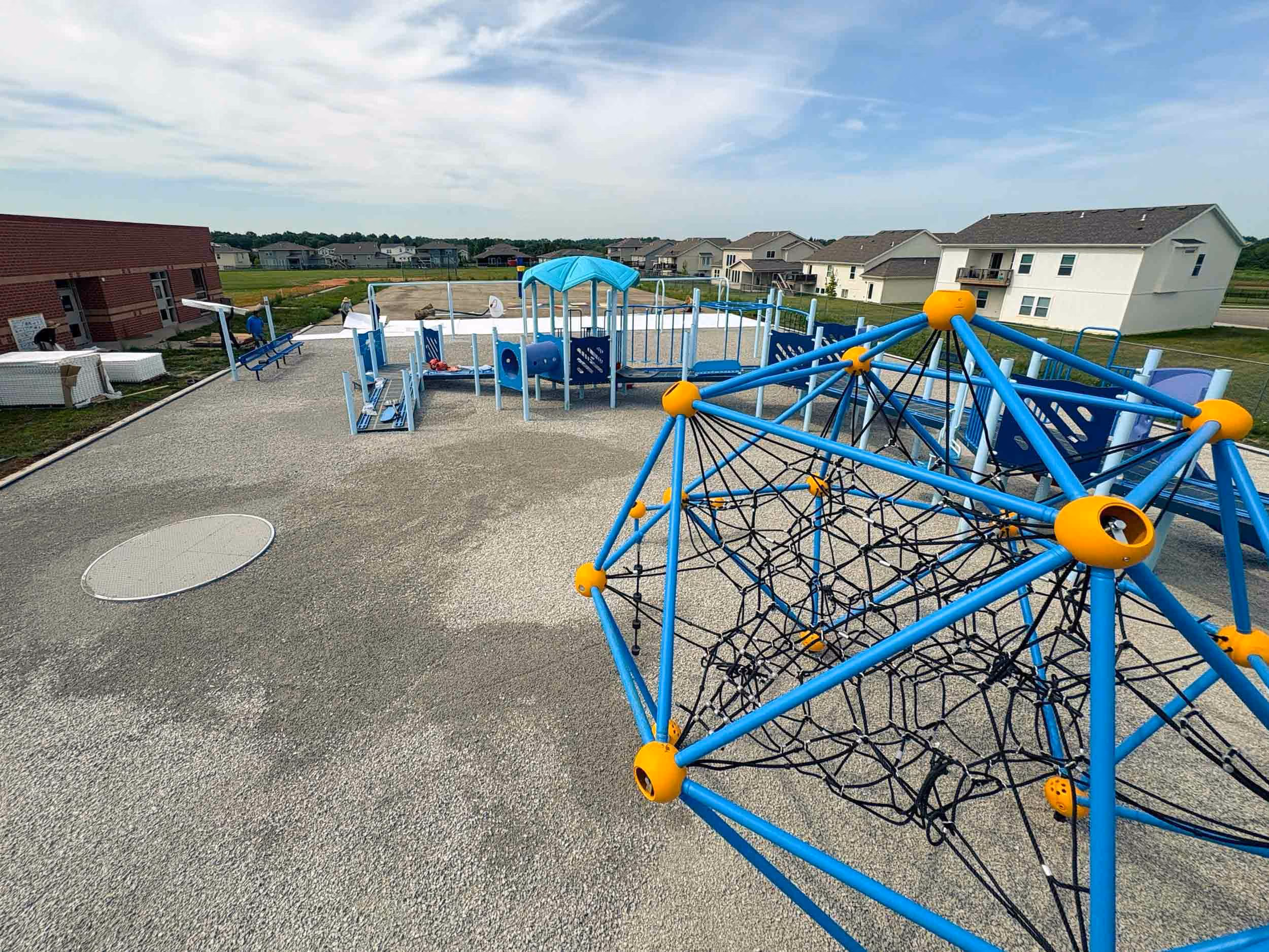 Outdoor playground with blue and yellow climbing structure, slides, and nearby houses under a partly cloudy sky.