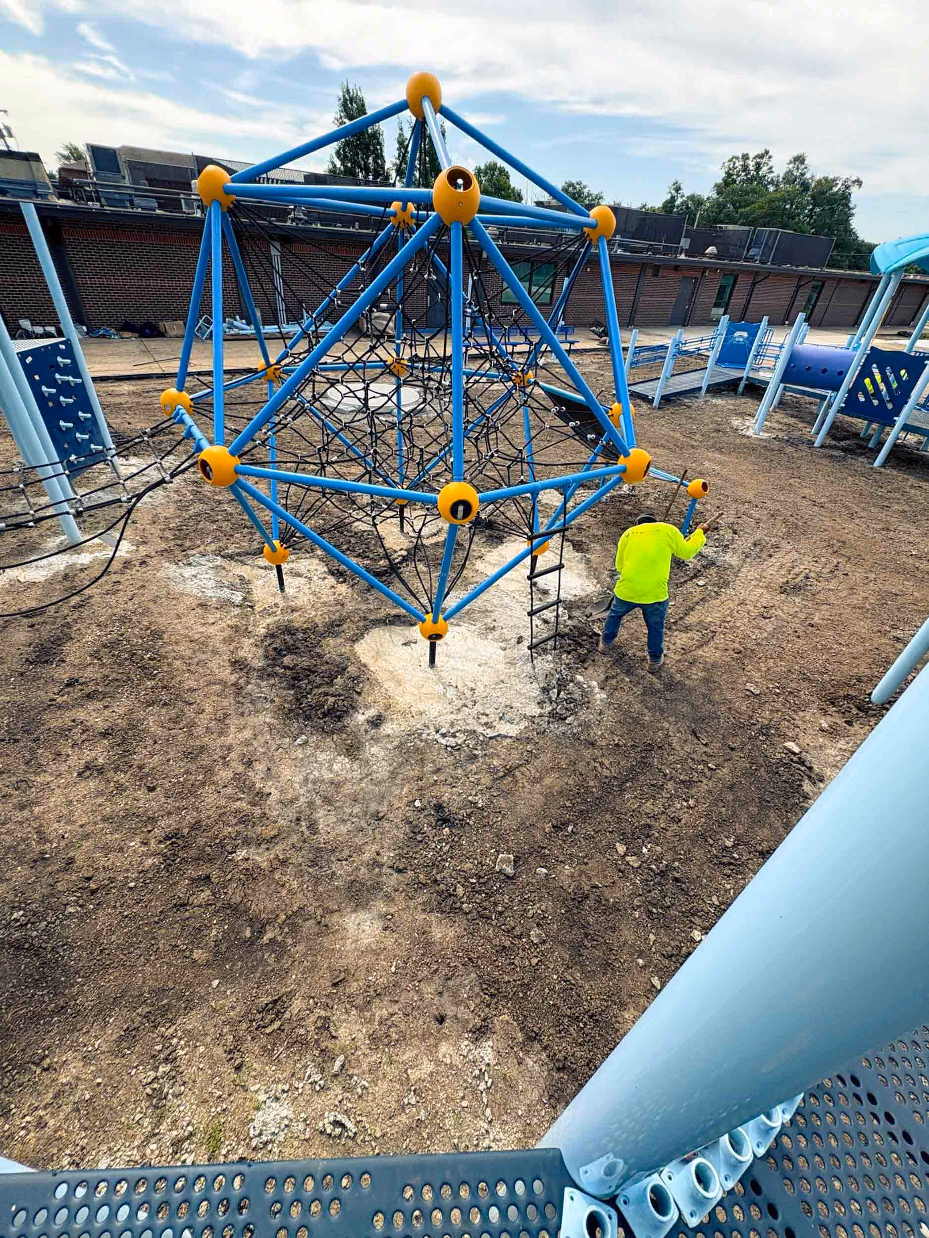 Worker in bright yellow jacket installs a blue and yellow rope climbing structure in playground under construction.
