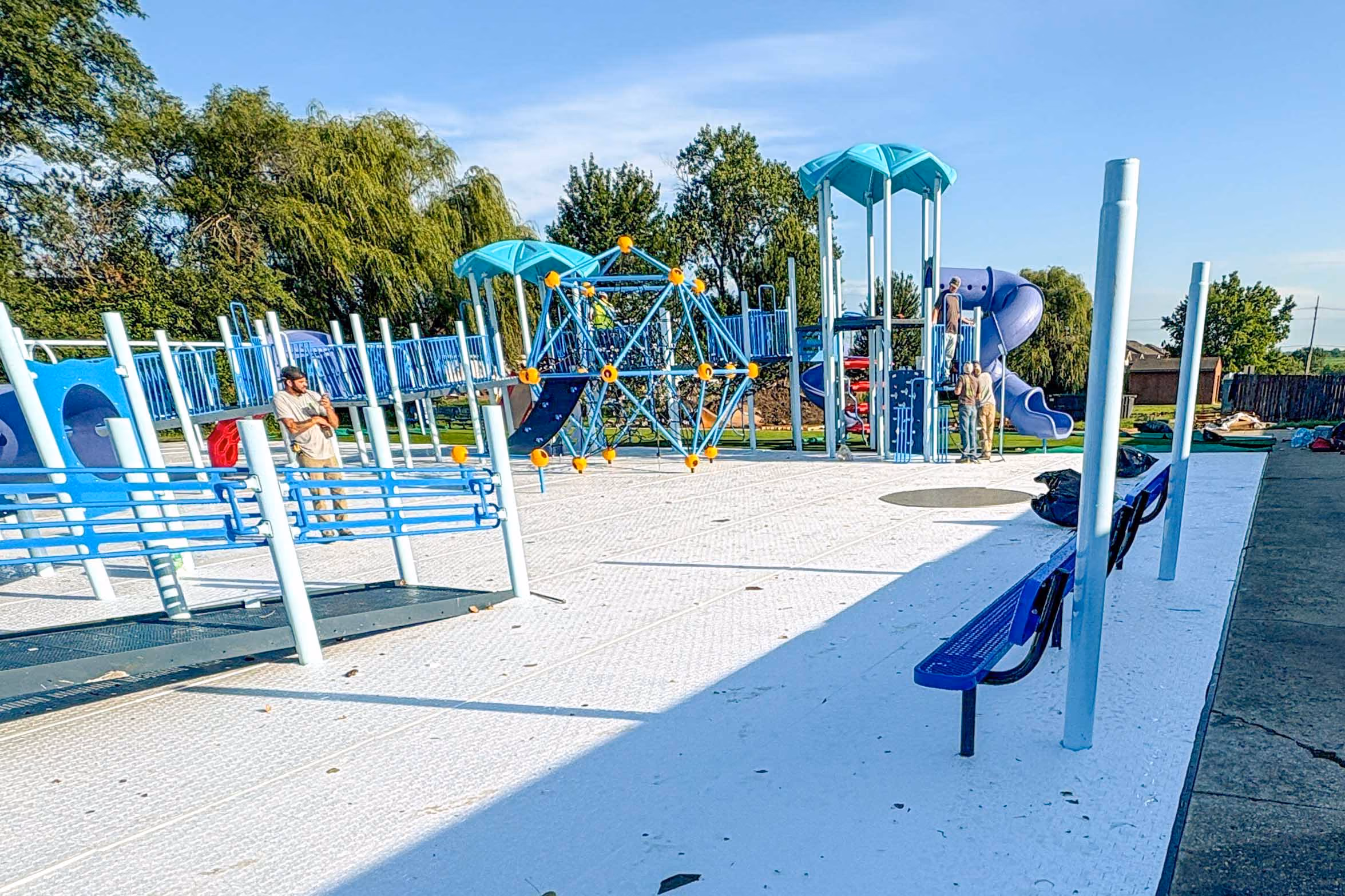 Playground with blue slides, climbing structures, and benches on a sunny day with trees in background.