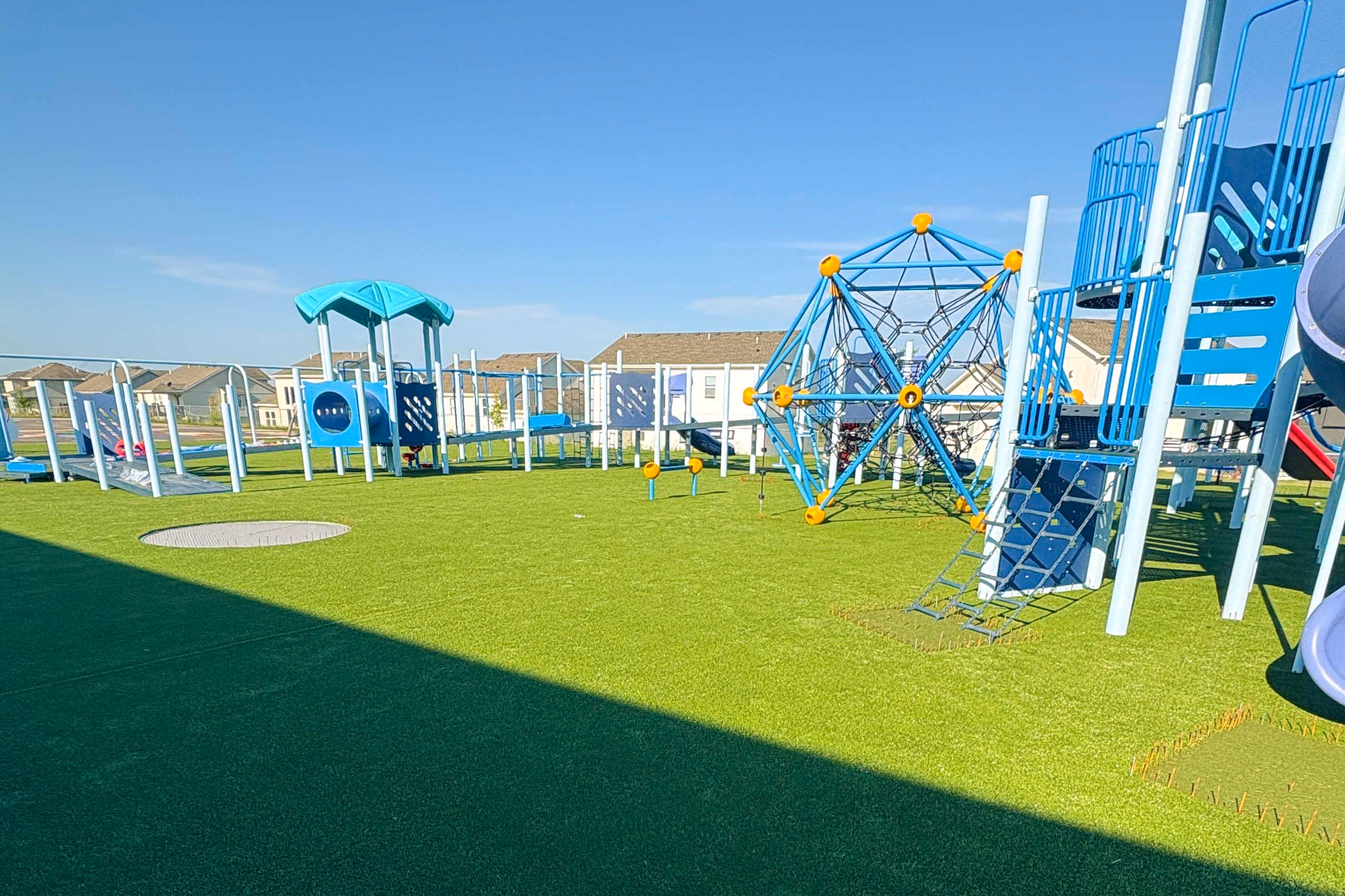 Empty modern playground with blue climbing structures, slides, and artificial grass under clear sky.