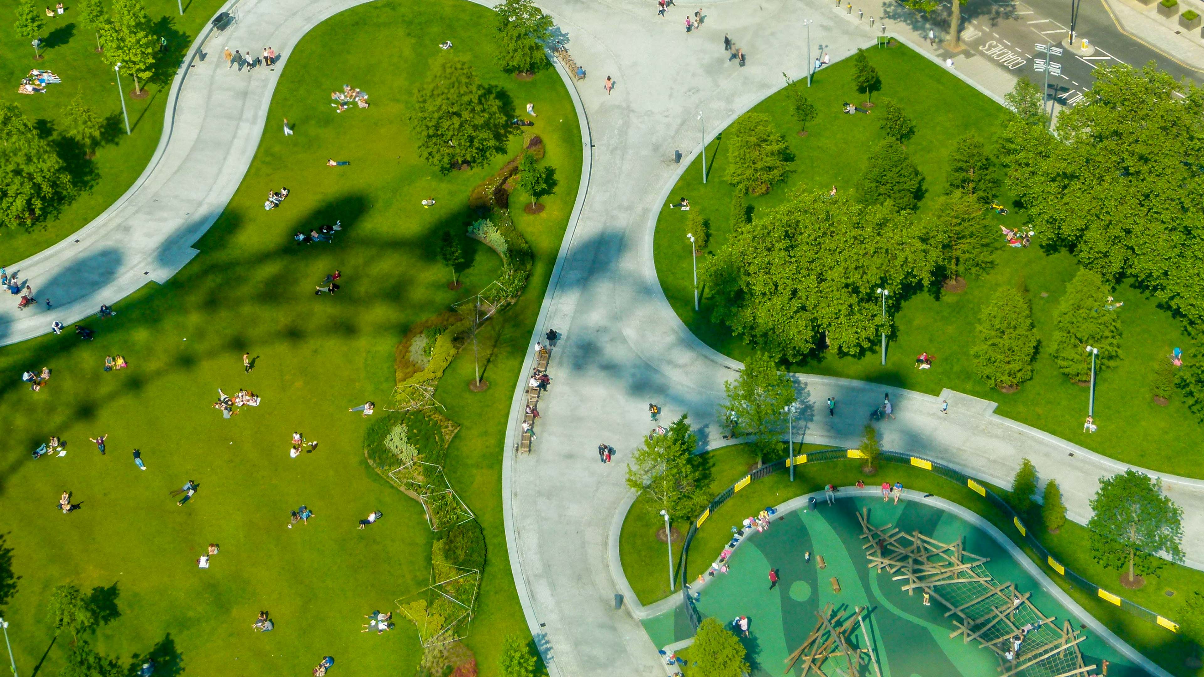 Aerial view of a sunny urban park with winding pale walkways, green lawns, scattered trees, people relaxing on the grass, and a playground in the lower right.