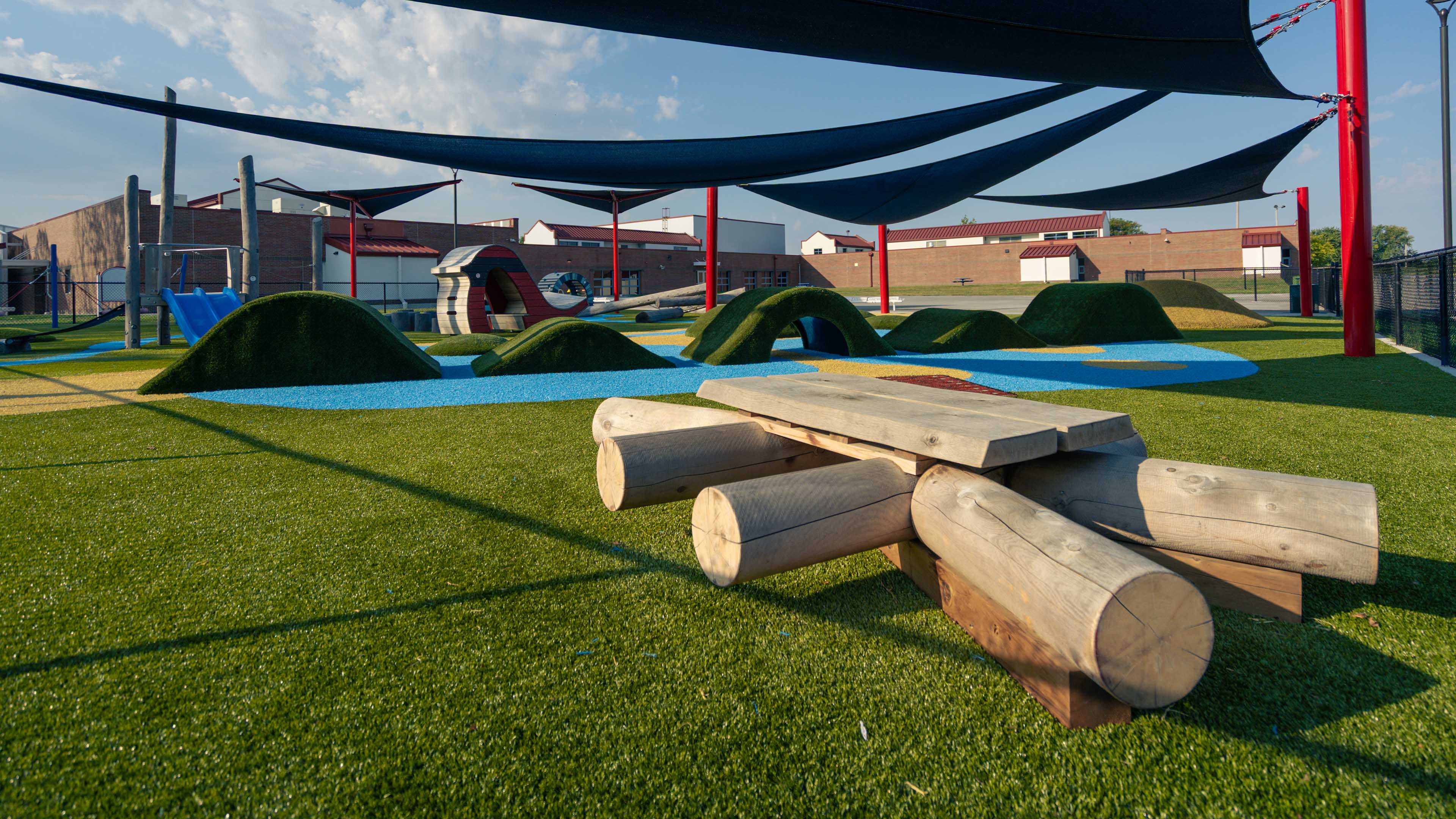An early childhood learning center playground with artificial turf, colorful safety surfacing, rolling turf mounds, wood play elements, slides, and large shade sails overhead.