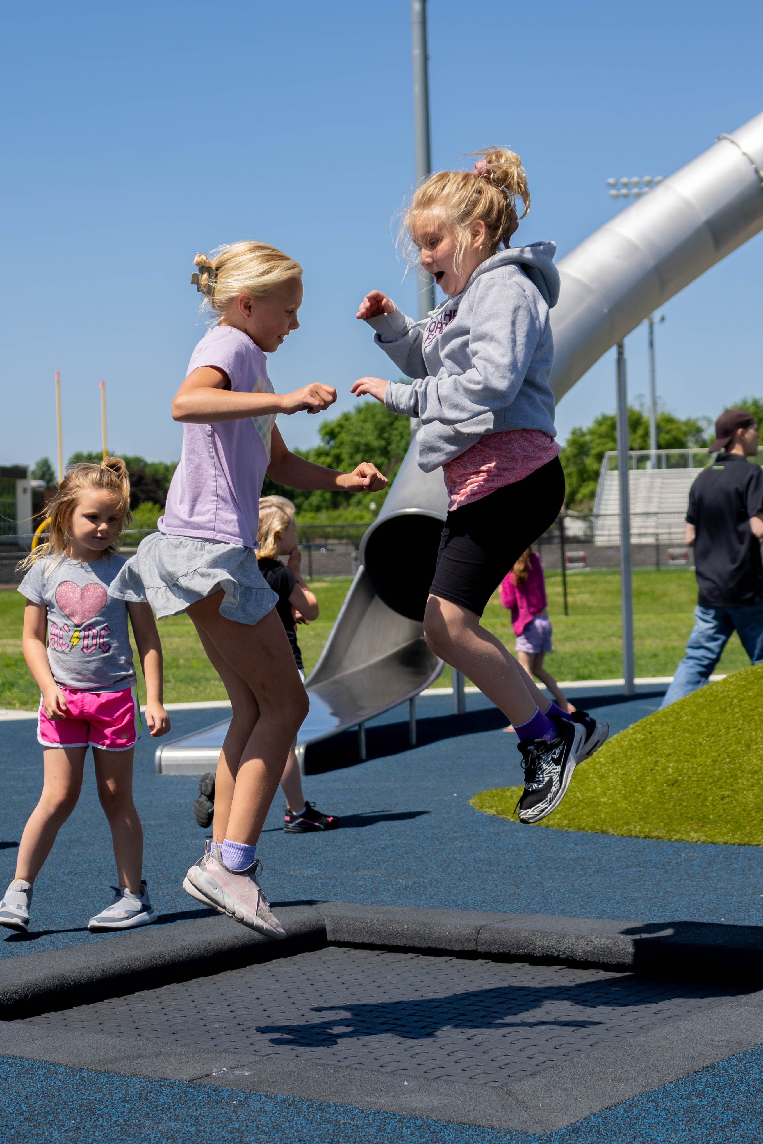 Children jump and play on a playground trampoline with a large metal slide and turf play mound in the background.
