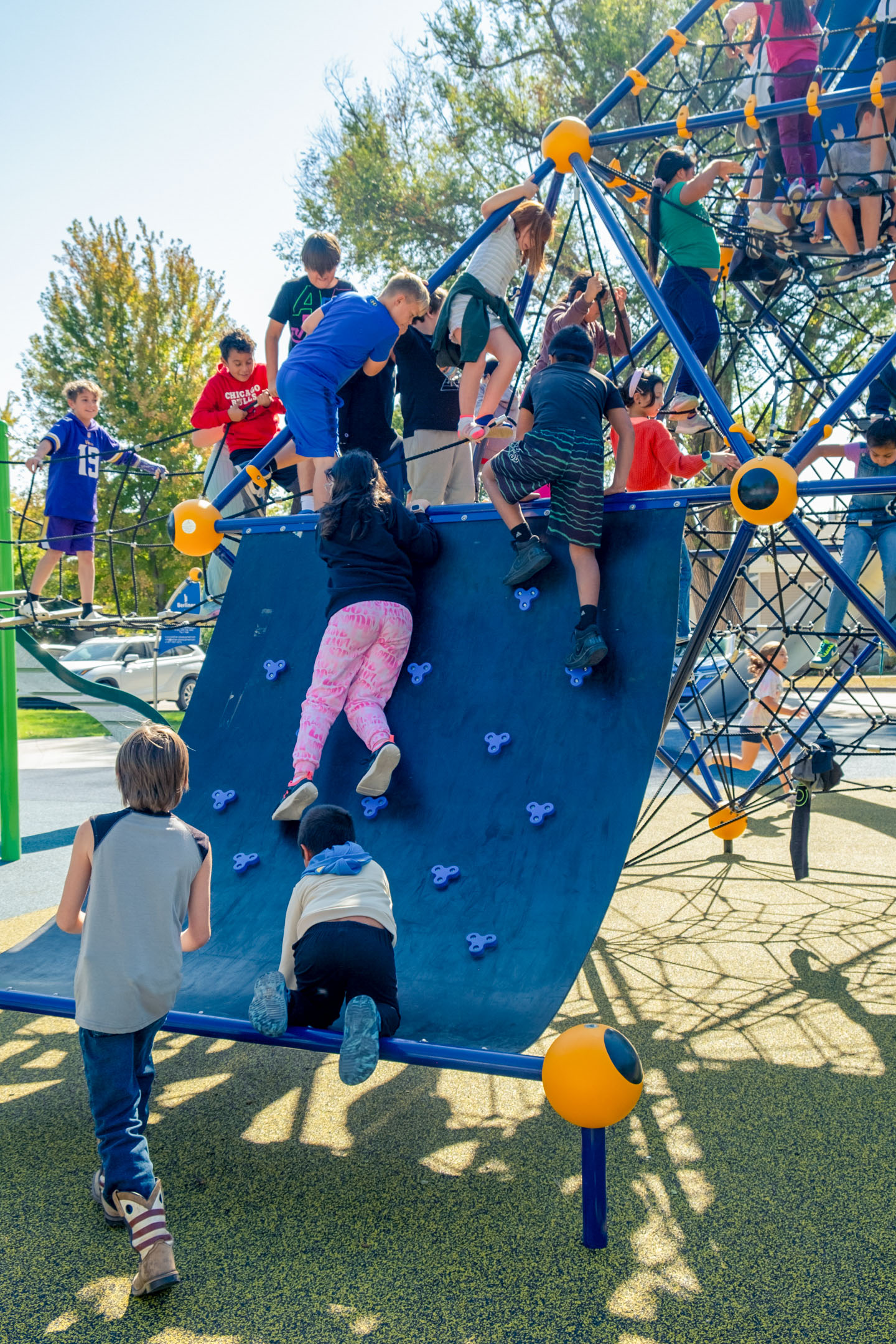 Children climb on a large playground rope structure with a curved climbing wall.