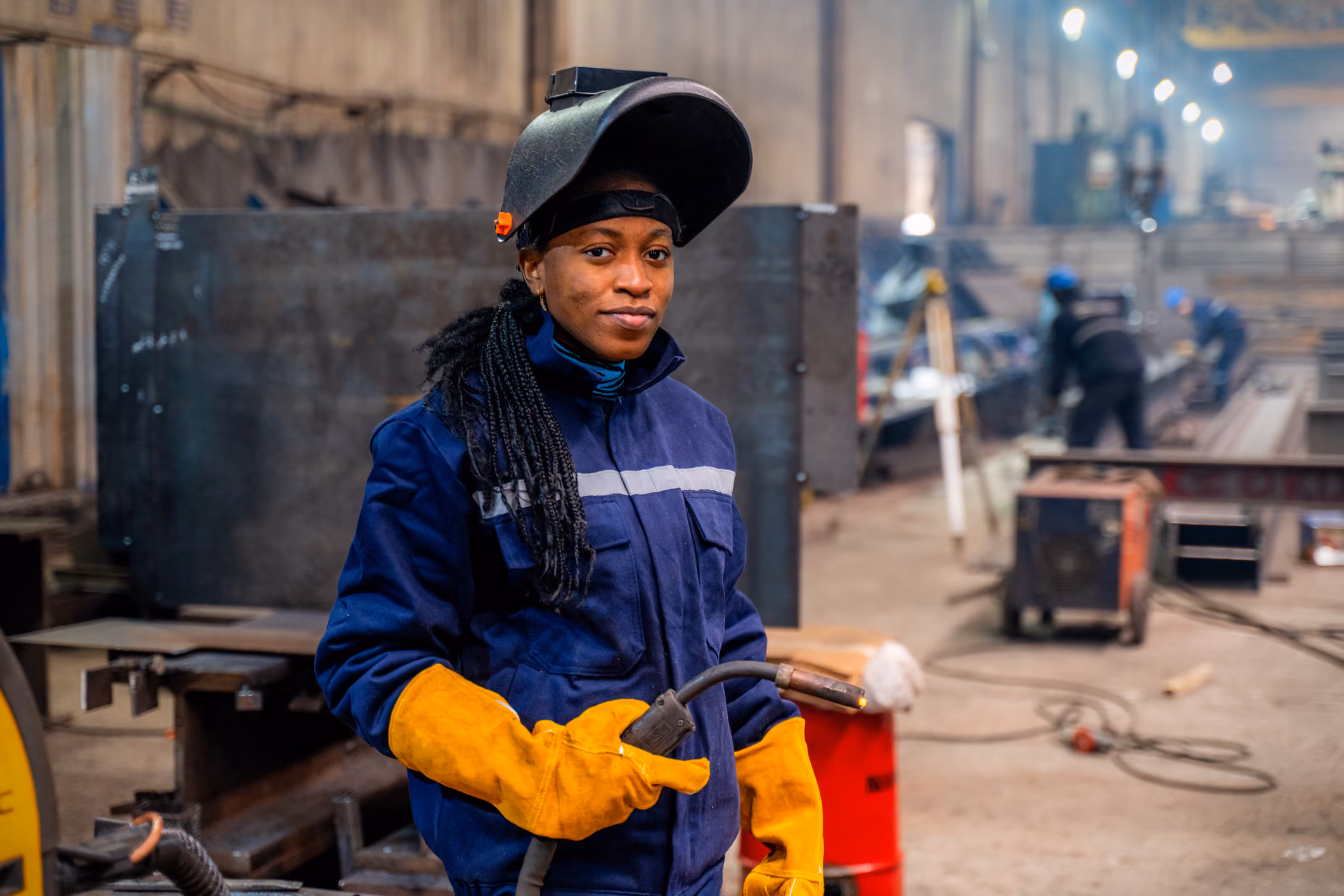 A woman in welding gear: mask, protective outfit, gloves, and welder.